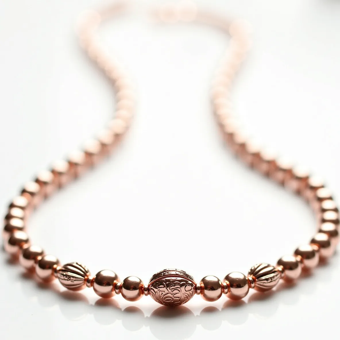 This rose gold necklace features a series of polished, spherical rose gold beads that create a smooth and elegant line. At the center, there is a larger oval bead with an intricate etched design that adds an element of sophistication and visual interest. Flanking this central bead are two other beads with a ribbed design, providing texture and contrast to the necklace's smooth components. The necklace is secured with a typical clasp, ensuring it stays securely fastened when worn. The combination of design elements makes this necklace both timeless and versatile for various occasions.