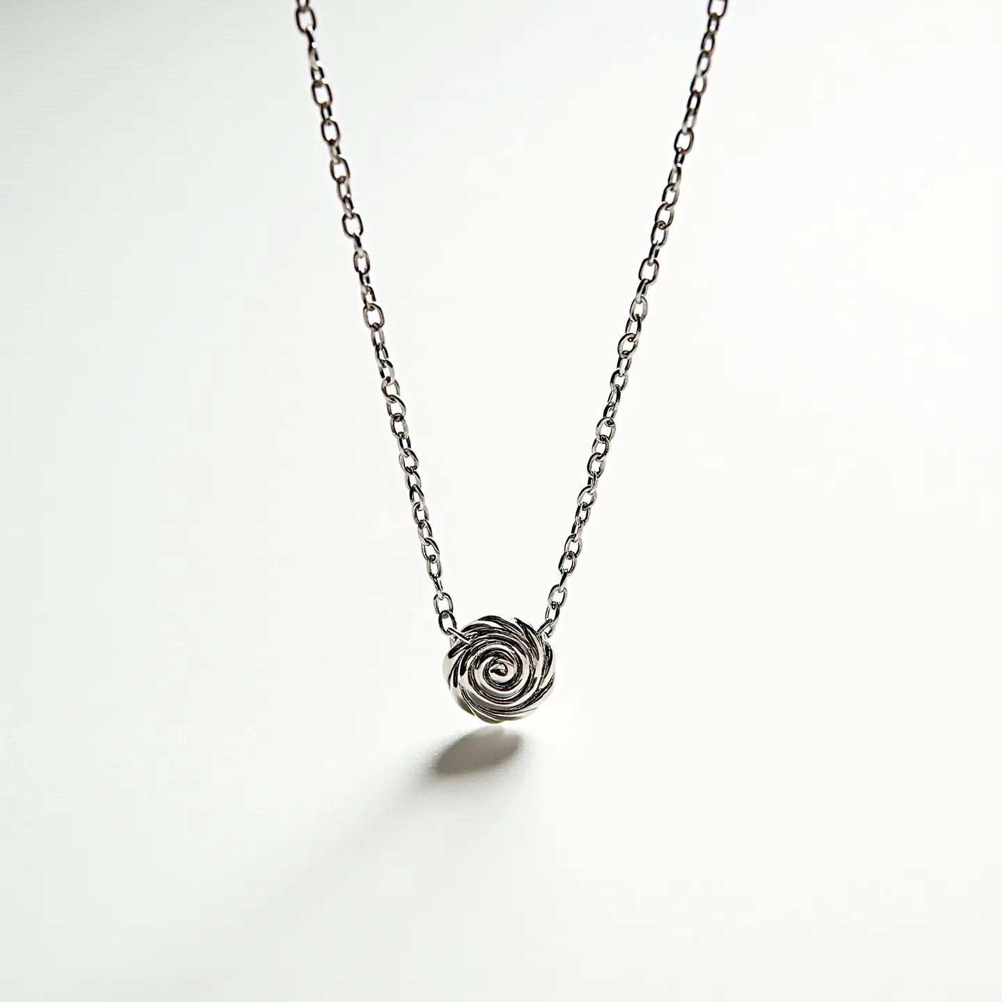 This rose necklace features a delicate pendant crafted in the shape of a rose, seemingly fashioned from a polished silver or similar metal, giving it a sleek and elegant appearance. The metal is formed into intricate loops that mimic the layered petals of a rose, showcasing fine craftsmanship. The pendant is affixed to a slender chain with interlocking links, likely made of the same material as the pendant, which contributes to its cohesive design. The necklace appears to include a simple yet functional clasp, ensuring easy wearability and secure attachment around the neck.