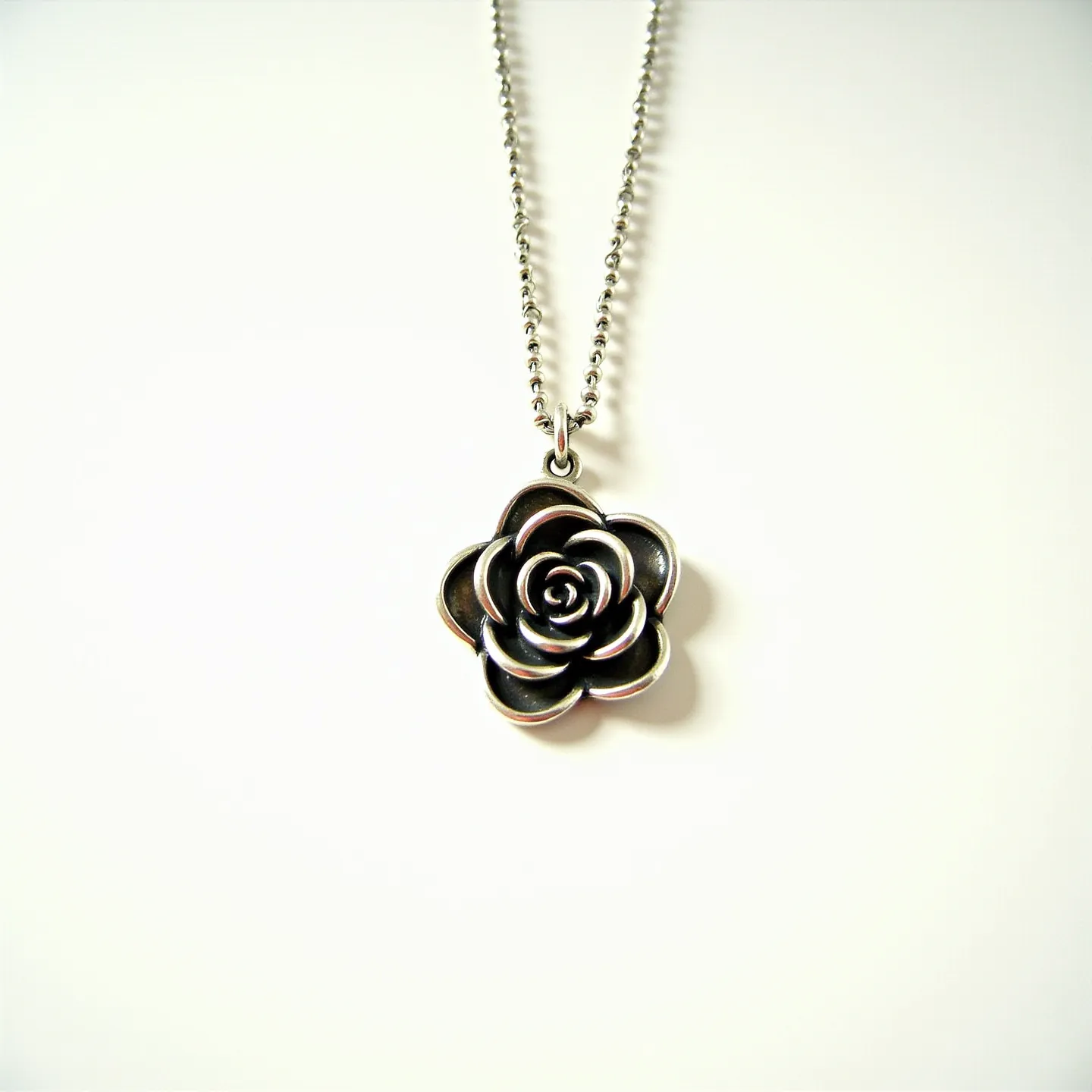 This rose necklace features a pendant shaped like a blooming rose, crafted from a metallic material with a silver tone, giving it a vintage-inspired appearance. The pendant exhibits intricate detailing that captures the layered petals of a natural rose. It is suspended from a delicate ball chain, also finished in a matching silver color, which complements the pendant's aesthetic. The necklace is equipped with a simple yet functional clasp, likely a lobster clasp, ensuring secure wearability. This elegant piece combines the organic beauty of a rose with the classic simplicity of understated jewelry design.