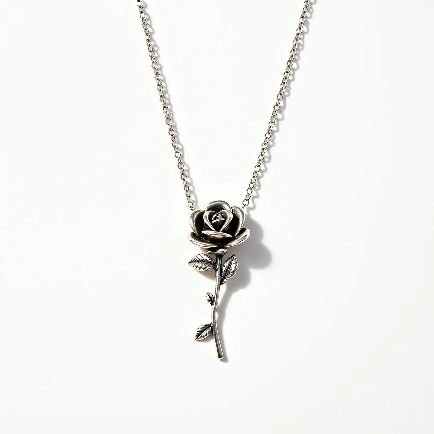 This rose necklace features a detailed design of a rose crafted from a metallic material, likely silver or a silver-toned metal, which gives it an elegant and classic appearance. The rose is depicted with precisely fashioned petals and leaves, enhancing its lifelike quality. The design showcases a combination of smooth and textured surfaces, creating a captivating play of light and shadow. The pendant hangs from a delicate chain with small, uniform links, contributing to its refined aesthetic. The chain is attached to the rose in a balanced manner, ensuring the pendant hangs securely. The necklace likely uses a standard clasp to fasten, maintaining the overall sleek and polished look.