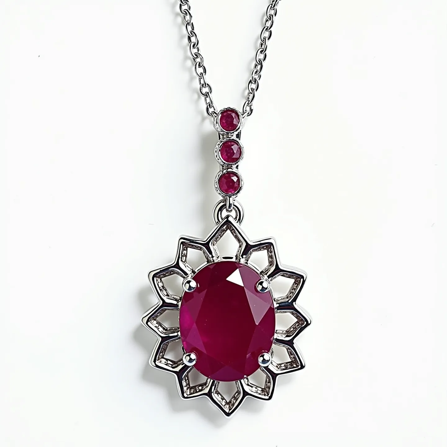 This ruby necklace features a striking design with a prominent oval-cut ruby set at its center. The ruby is secured with four prongs and encircled by a starburst pattern crafted from a metal resembling polished silver. Above the main ruby, the necklace includes a series of three small round-cut rubies, each set in a bezel setting, adding a graceful vertical accent to the design. The chain appears to be made of a similar polished metal, linking seamlessly to the pendant with a sturdy attachment, ensuring a cohesive and elegant presentation.