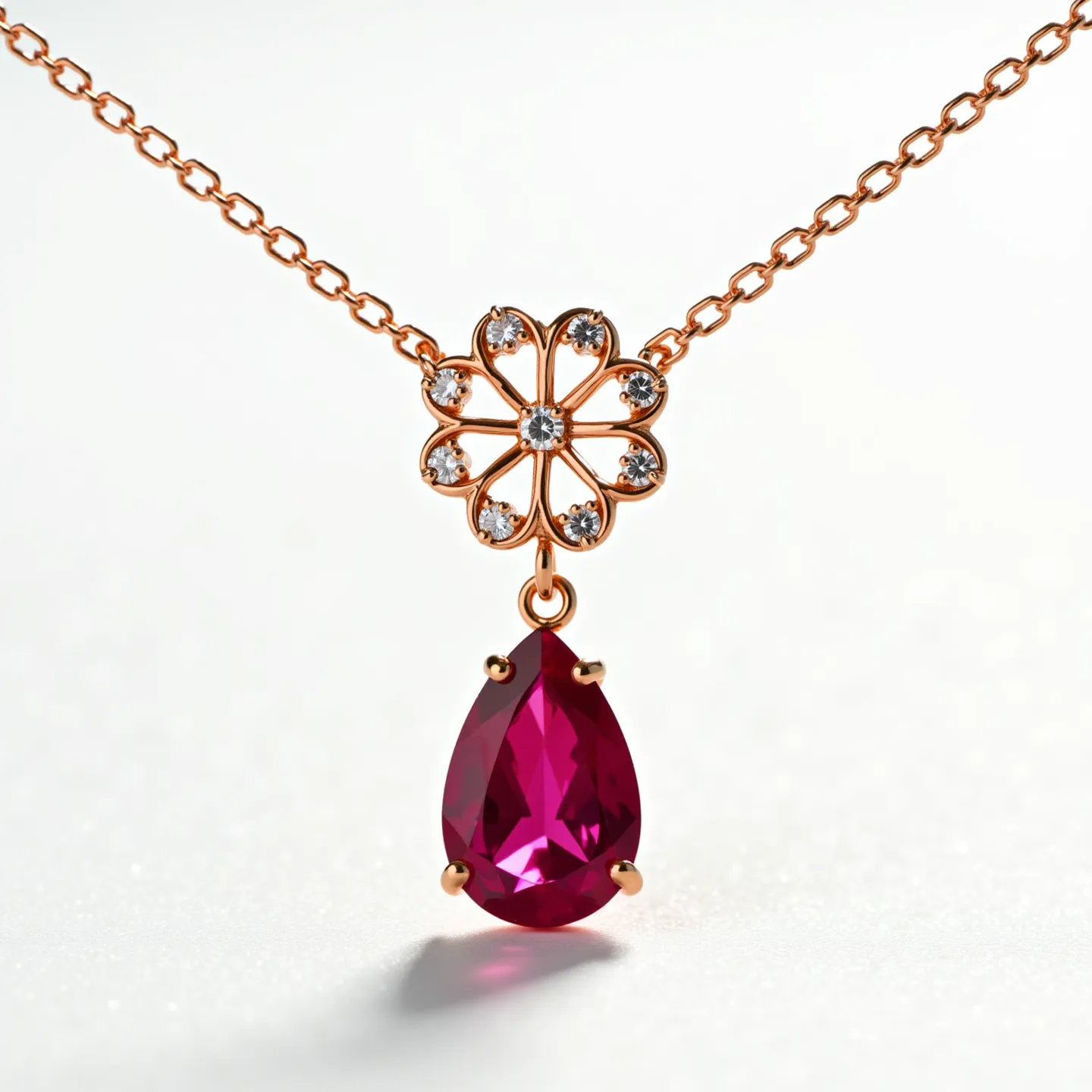 This ruby necklace features a striking pear-cut ruby pendant set in a delicate prong setting, creating a focal point with its deep red hue. The pendant is suspended from a chain made of what appears to be a rose gold material, lending a warm, luxurious tone to the piece. Above the ruby, a flower-shaped motif is adorned with small, sparkling gems, likely diamonds, set in a bezel style to complement the central stone. The necklace showcases an elegant chain link design that likely includes a clasp, ensuring a secure and comfortable fit when worn.