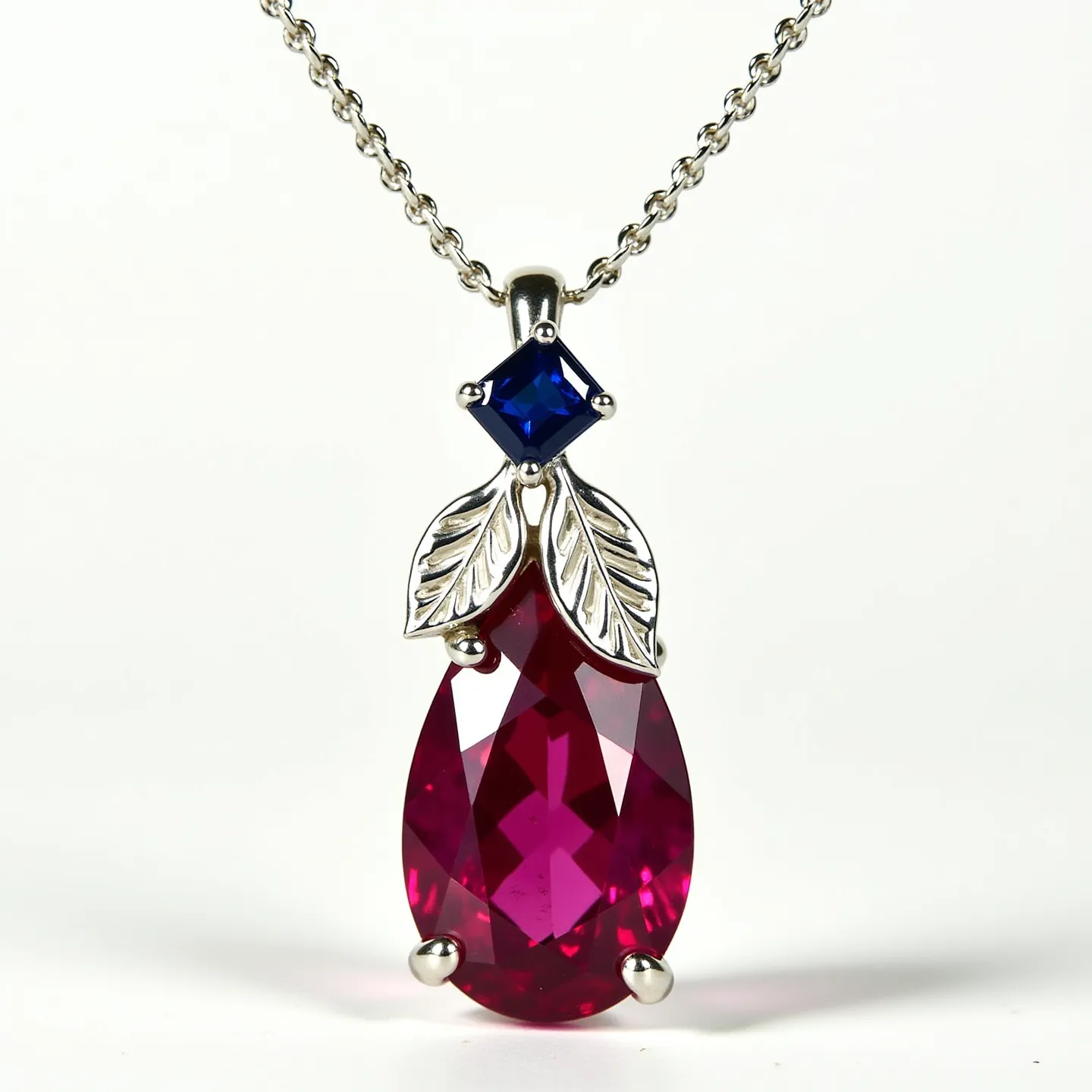 This ruby necklace features a stunning pear-shaped ruby, cut to showcase its vibrant red hue, set in a minimalist prong setting. Above the ruby, a square-cut blue gemstone, possibly a sapphire, is secured in a similar prong setting, adding a striking contrast to the design. The necklace is accented with delicate metalwork, resembling two leaves, crafted from silver-toned metal, enhancing the natural elegance of the piece. It is suspended from a fine, silver-colored chain that completes the sophisticated look, with a simple and functional clasp that ensures secure wear.