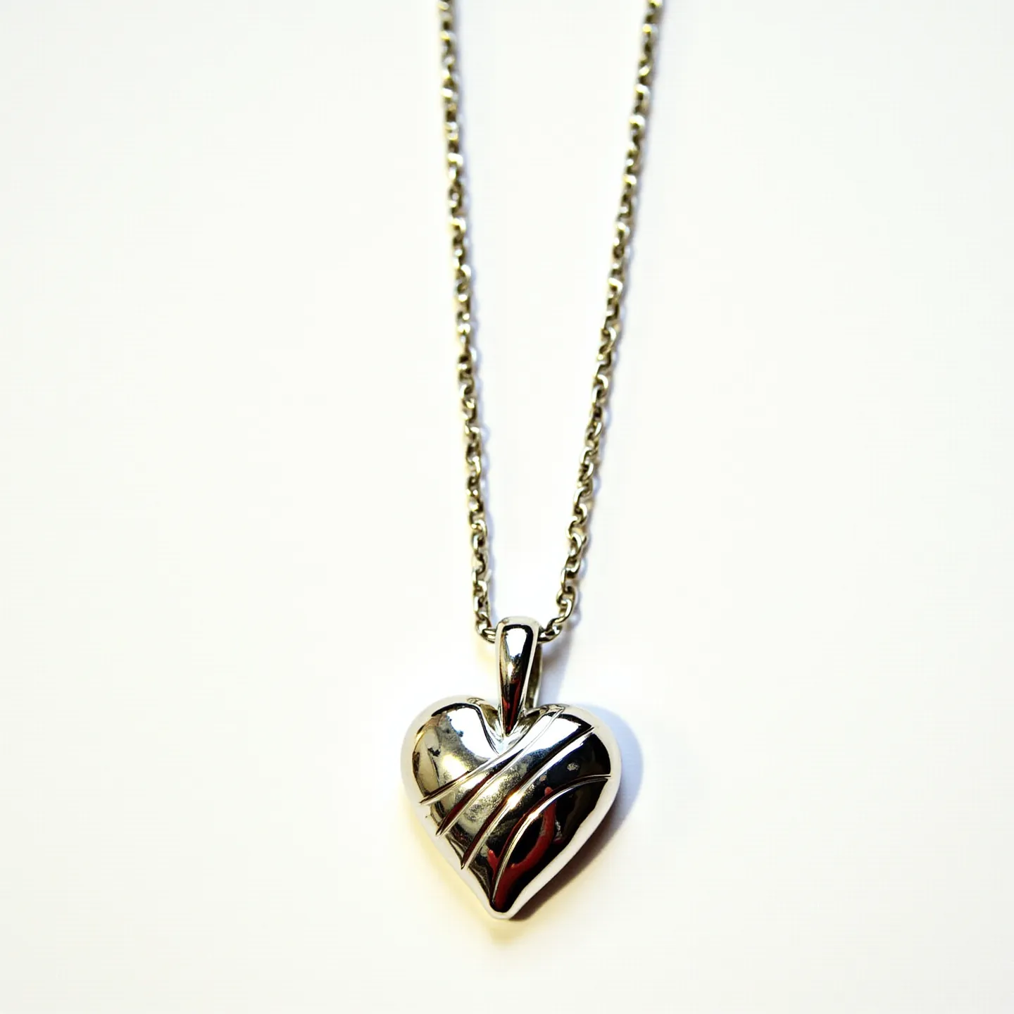 This sacred heart necklace features a polished silver heart-shaped pendant with an intricate design that resembles wrapped banding across its surface. The pendant seems to have a smoothly contoured finish, likely crafted from sterling silver, displaying a shiny and reflective quality. The necklace chain is composed of interlocking links, giving it a durable and classic appearance, possibly made from the same metal as the pendant for a cohesive look. The pendant is attached to the chain via a looped bail that allows for smooth movement along the chain. There are no evident gems or stones set in the pendant, and the chain likely has a standard clasp for secure attachment around the neck.