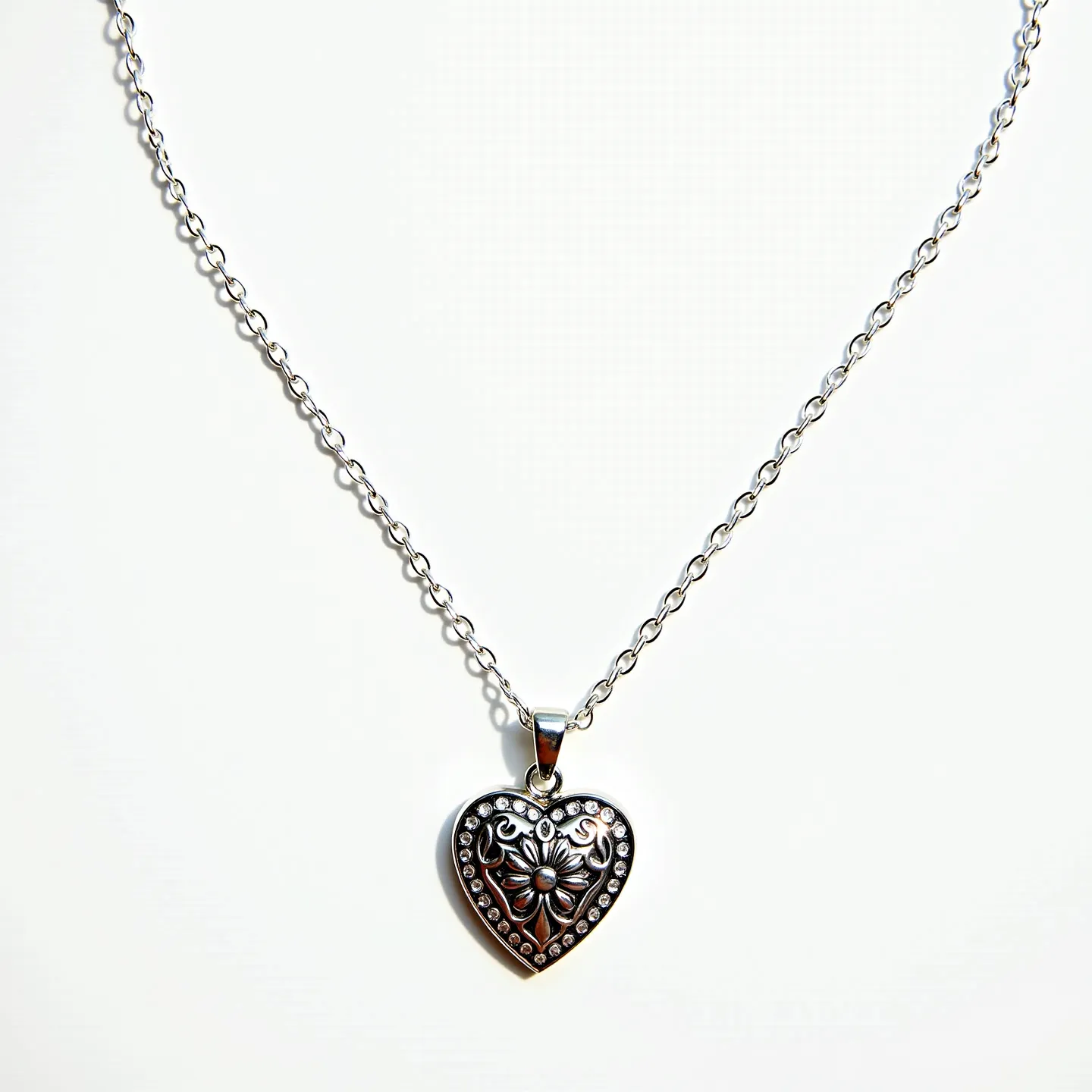 This sacred heart necklace features a heart-shaped pendant crafted from a silver-toned metal with ornate detailing, showcasing a central floral design surrounded by a border of small, clear stones, likely set into the metal to create a sparkling effect. The stones appear to be round cut, adding a touch of elegance to the intricate pendant. The pendant is suspended from a polished metal bail, connecting to a simple chain with interconnected links, providing a classic and timeless appearance. The necklace is fastened with a standard clasp, ensuring secure and comfortable wear.