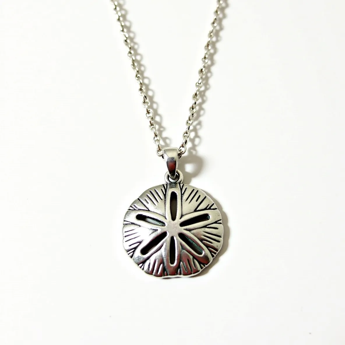 This sand dollar necklace features a beautifully crafted pendant resembling a sand dollar, likely made from sterling silver given its metallic sheen and intricately detailed design. The pendant showcases a combination of polished and oxidized finishes to highlight the natural patterns of a sand dollar. It hangs from a sturdy, matching silver chain, lending a cohesive aesthetic to the entire piece. The necklace is secured with a classic lobster clasp, allowing for easy wearing and removal. The pendant's loop attachment is seamlessly integrated into the design, maintaining the necklace's elegant and sophisticated appearance.