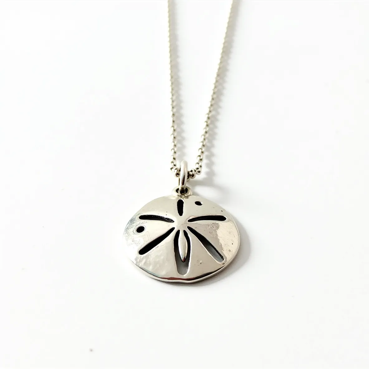 This sand dollar necklace features a beautifully crafted pendant shaped like a sand dollar, likely made from polished silver, giving it a shiny and elegant appearance. The pendant showcases intricate cut-out designs mimicking the natural pattern of a sand dollar. It is attached to a delicate silver chain that complements the pendant’s metallic finish. The chain appears to have a simple loop clasp mechanism, which ensures secure wear while maintaining the necklace's elegant simplicity. The pendant and chain work together to create a cohesive and sophisticated piece of jewelry.