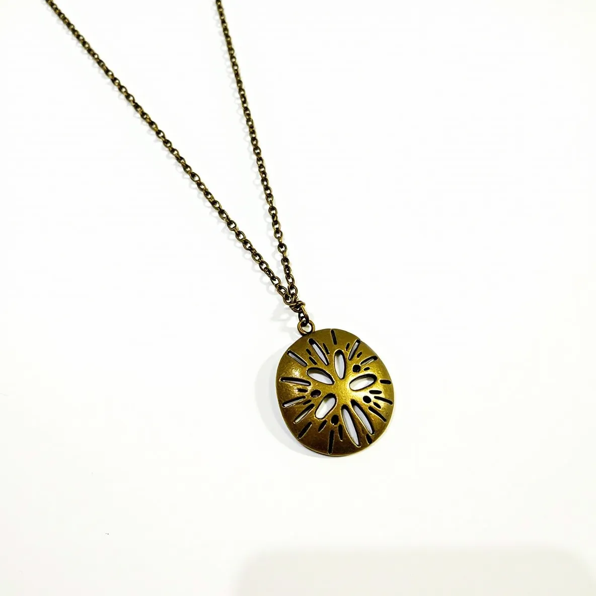 This sand dollar necklace features a pendant crafted from a bronze-toned metal with intricate cutouts mimicking the natural design of a sand dollar. The pendant hangs from a matching chain composed of interlinked metal loops, which adds a rustic charm to the overall appearance. Connected by a simple, unobtrusive ring, the pendant seamlessly dangles from the chain, allowing for ease of movement. The necklace appears to be fastened with a lobster claw clasp, providing secure wearability. The design does not include any gemstones, focusing solely on the elegant metalwork to capture attention.