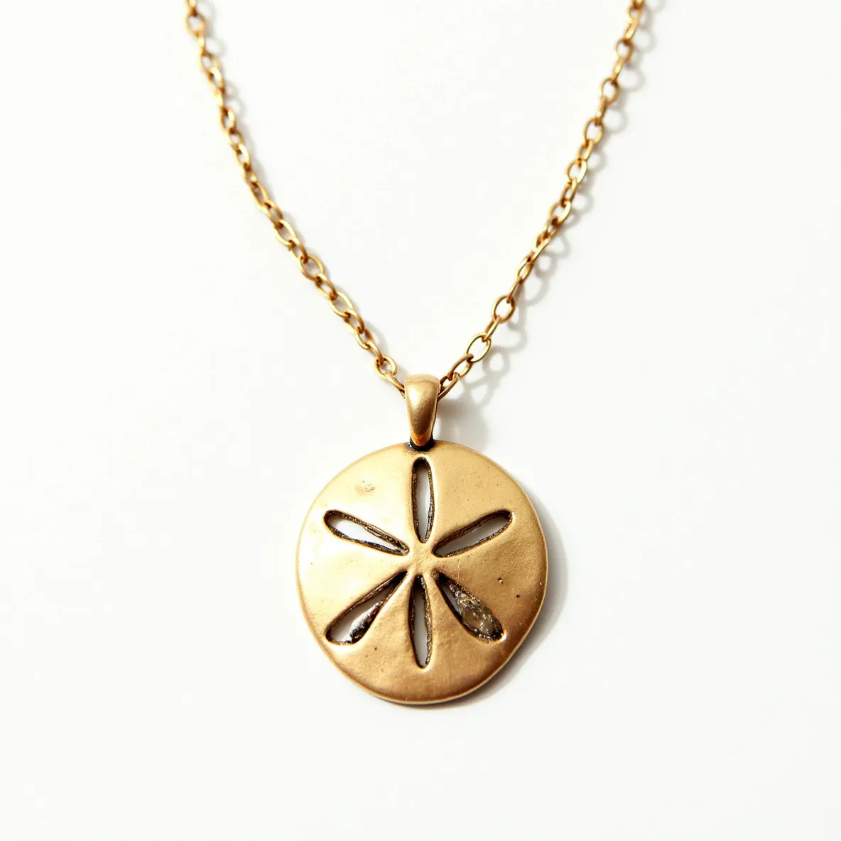 This sand dollar necklace features a round pendant designed to resemble a sand dollar, made from a gold-toned metal. The pendant is intricately crafted with five cut-out slots, characteristic of a sand dollar design, and is attached to a delicate chain. The chain itself consists of small, uniform links, matching the gold tone of the pendant. A simple clasp attaches the necklace securely. This elegant piece does not incorporate any gemstones, focusing instead on the distinctive sand dollar motif.