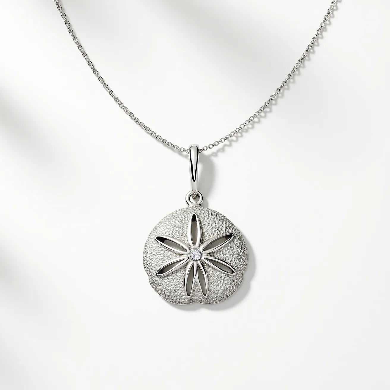 This sand dollar necklace features a detailed, metallic sand dollar pendant, crafted in a polished metal that resembles silver, emphasizing its intricate texture. At the center of the sand dollar is a small, round gemstone, likely a diamond, cut to sparkle and set in the middle to enhance the natural design. The pendant is suspended from a delicate chain, possibly in a matching metal, ensuring a cohesive look. The attachment mechanism includes a sleek bail that holds the pendant securely to the chain, allowing fluid movement while worn.