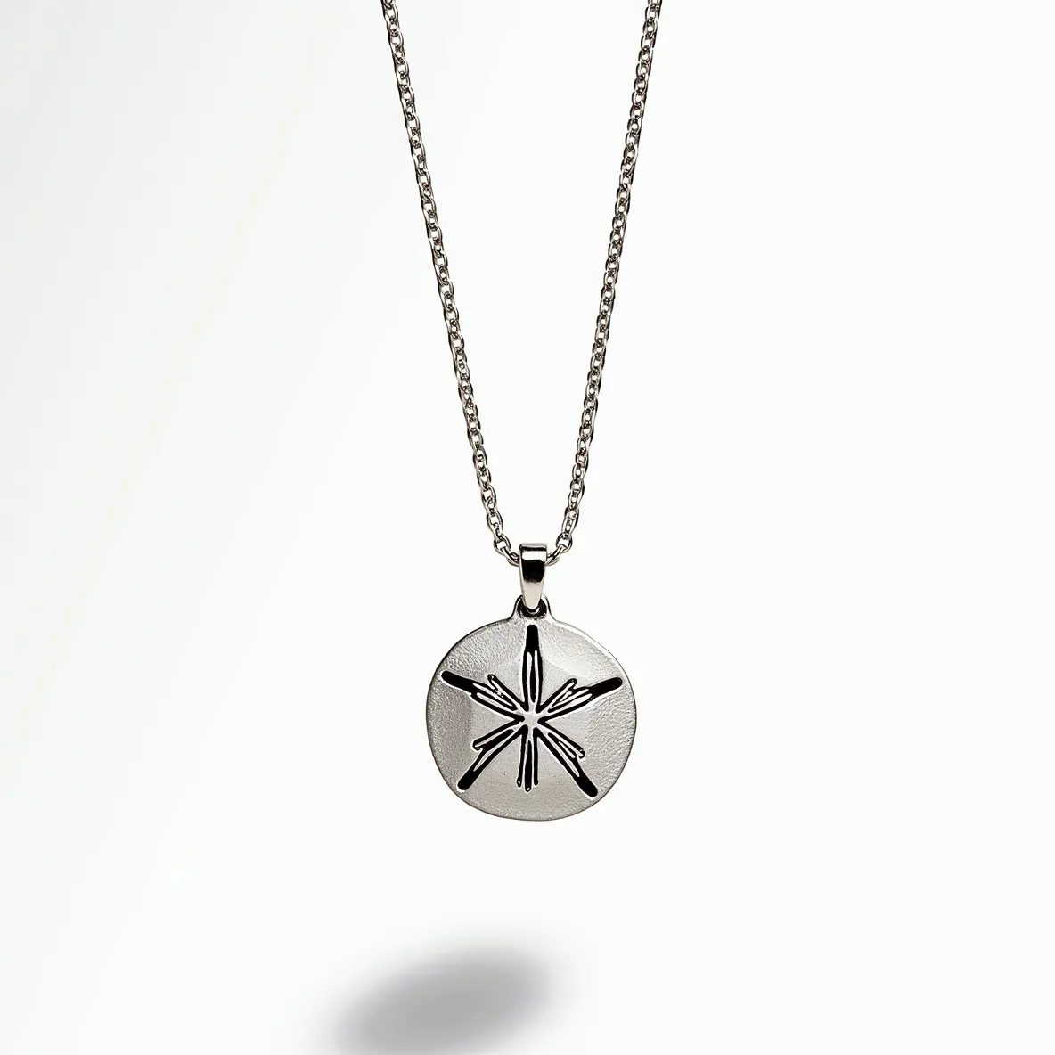 This sand dollar necklace features a silver-toned metal pendant with a detailed sand dollar design. The pendant is smoothly polished and casts a subtle shine, emphasizing the intricate star-shaped pattern at its center. It is suspended from a fine, complementary chain, likely made of the same metal, which adds to the elegance of the piece. The chain is equipped with a simple but secure clasp that ensures ease of wear. The overall design is minimalist yet stylish, capturing the essence of beach-inspired jewelry.