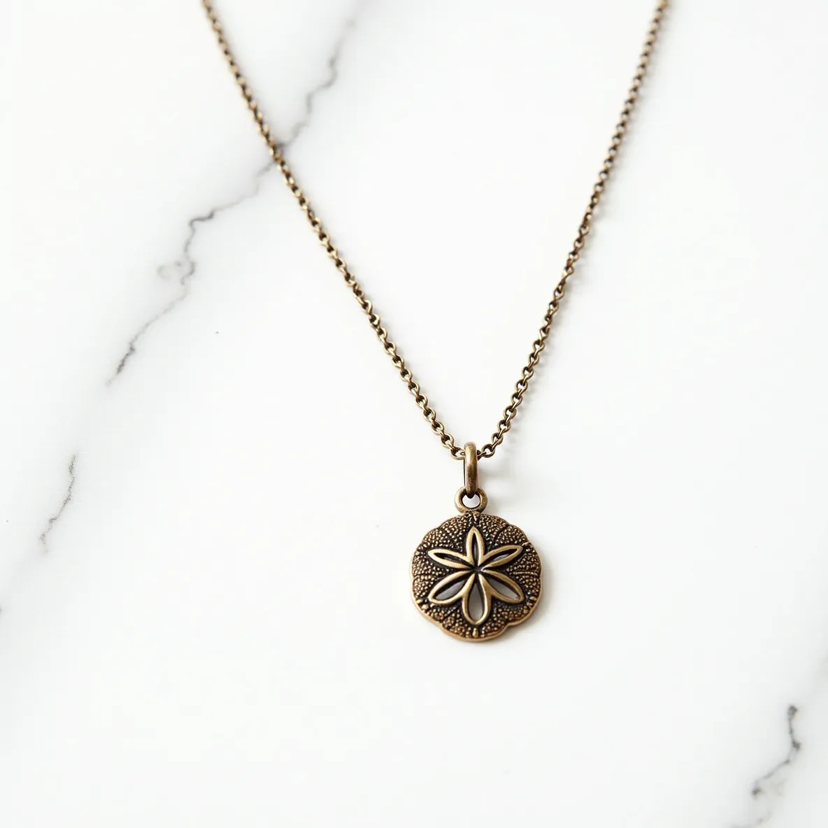 This sand dollar necklace features a pendant crafted from a metal with a bronze-like finish, giving it a rustic and vintage appearance. The sand dollar pendant showcases a delicate, embossed star pattern in the center, adding a touch of elegance to the design. Suspended from a finely linked chain, the pendant is secured to the necklace with a simple bail that ensures stability and free movement. The necklace does not appear to include any gemstones, focusing instead on the intricate metalwork of the sand dollar itself. The chain likely closes with a standard clasp, completing the subtle, seaside-inspired aesthetic of the piece.