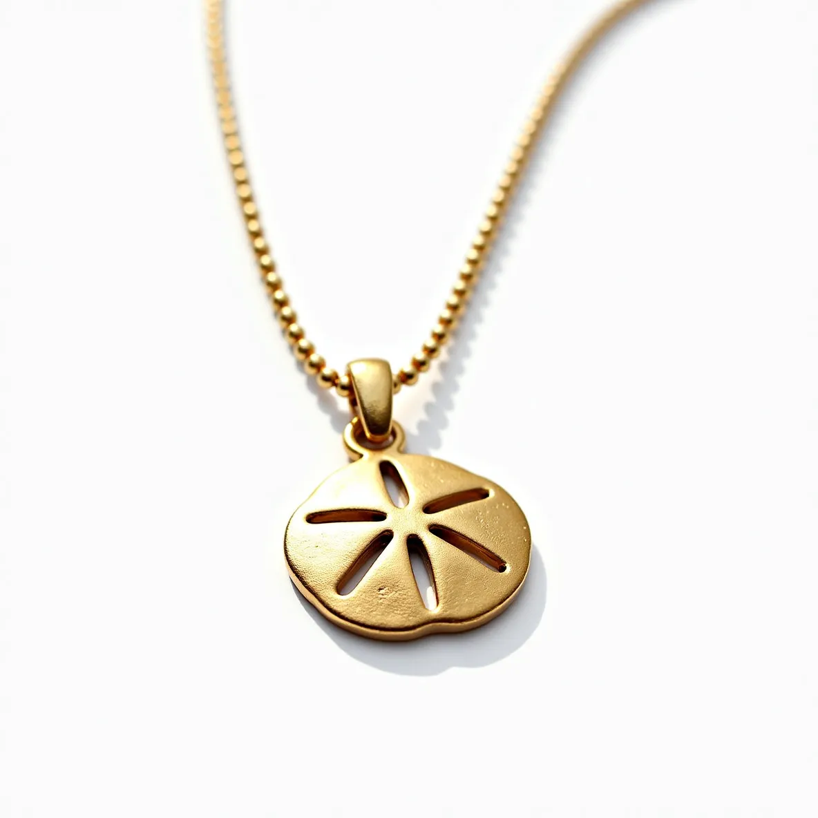 This sand dollar necklace features a polished gold or gold-toned metal pendant designed in the shape of a sand dollar, with finely crafted cut-out details that mimic the natural markings of the sea creature. The pendant is attached to a matching gold or gold-toned chain with small, evenly spaced links, lending a cohesive and elegant appearance. The attachment mechanism is a smooth, rounded bail that connects the pendant to the chain seamlessly. The overall design is minimalist yet sophisticated, highlighting the delicate beauty of the sand dollar form in a warm metallic finish.