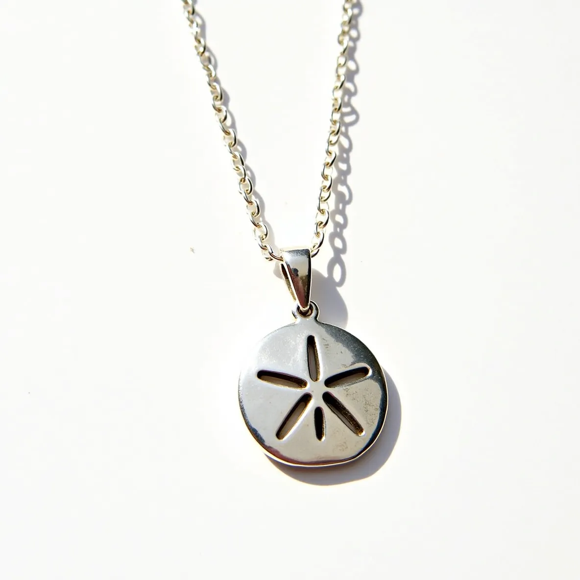 This sand dollar necklace features a polished metal pendant crafted in the shape of a sand dollar, showcasing intricate cut-out details that mimic the natural design of a real sand dollar. The pendant is suspended from a delicate chain, likely made of a similar metal, indicating a cohesive and elegant design. The attachment is a simple loop connected to a classic chain link, ensuring secure and comfortable wear. The entire piece highlights a minimalist aesthetic, focusing on the natural beauty and symbolism of the sand dollar.