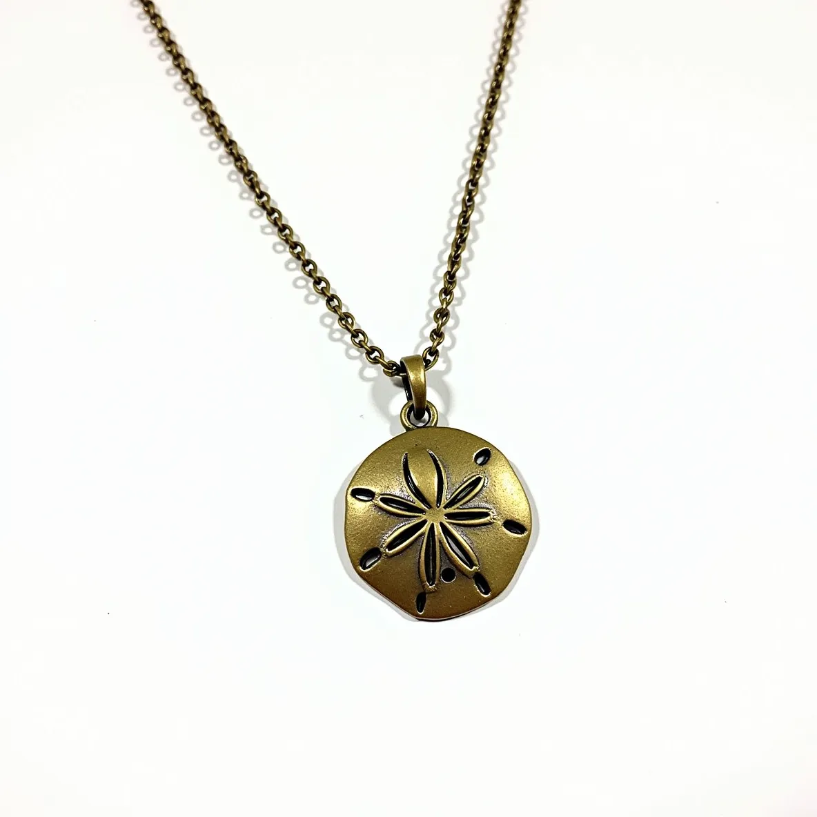 This sand dollar necklace features a round pendant crafted from a metal with a brass or bronze finish, giving it a warm, earthy tone. The pendant displays delicate carvings that mimic the natural design of a sand dollar, with symmetrical cutouts forming a star pattern at its center. The chain appears to be made of the same metal, composed of small interlinked loops that provide a classic, understated look. The pendant is attached to the chain via a simple metal loop that complements its design, ensuring a seamless look between the chain and pendant. This necklace does not include any gemstones, focusing instead on the intricately shaped metalwork that captures the essence of a sand dollar.