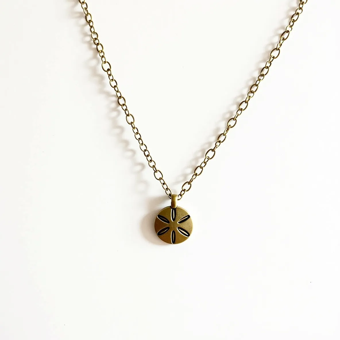 This sand dollar necklace features a delicate chain, likely made from metal with an antique bronze finish, evoking a vintage look. The pendant itself is shaped like a sand dollar and maintains a similar bronze hue to match the chain, with engraved details to mimic the natural design of a sand dollar. The pendant is attached via a simple loop connector that aligns seamlessly with the chain. There are no visible gems or stones incorporated into the design, and the necklace is closed with a standard lobster clasp that provides a secure and adjustable fit.