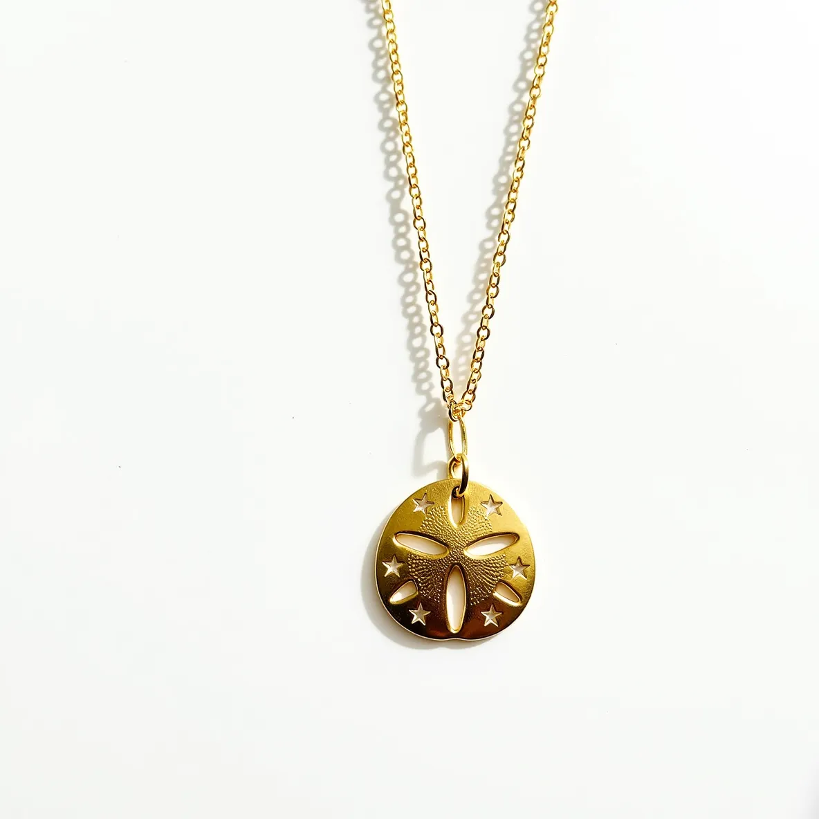 This sand dollar necklace features a beautifully crafted pendant made from gold-toned metal, showcasing intricate cutout details that mimic the natural patterns of a sand dollar. The pendant is adorned with small, star-shaped embellishments that add a touch of whimsy and elegance. The necklace is attached to a delicate chain, also in a matching gold tone, which complements the pendant nicely. It includes a standard jump ring that links the chain to the pendant, allowing for smooth and secure wear.