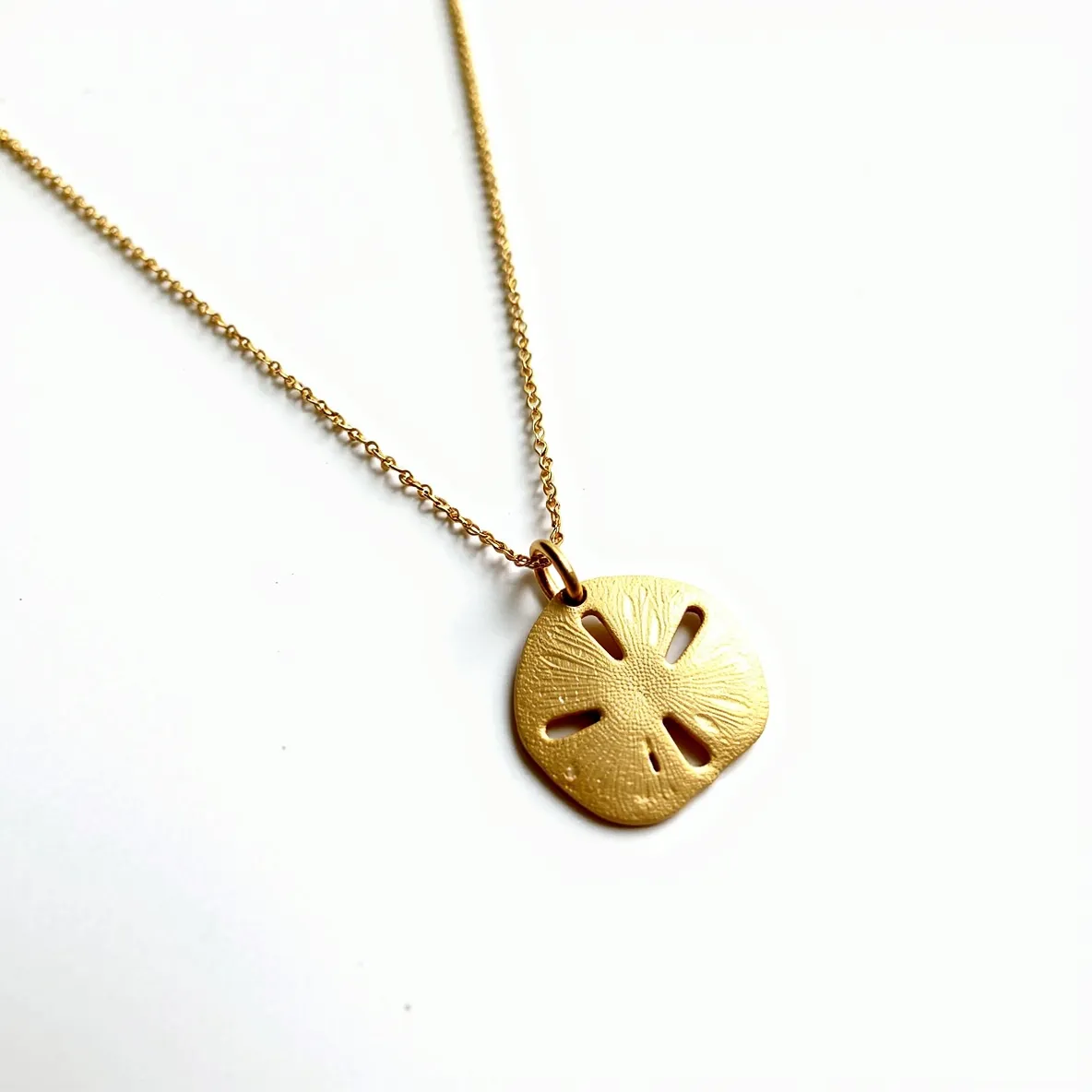 This sand dollar necklace features a pendant crafted from a material with a rich, golden hue, evoking the elegance of gold or a gold-tone finish. The pendant is designed with intricate details that resemble the natural texture of a real sand dollar, contributing to its elegance and authenticity. It hangs from a delicate chain, likely made of a matching metallic material, enhancing the cohesive look of the piece. The necklace appears to include a small loop, seamlessly attaching the pendant to the chain.