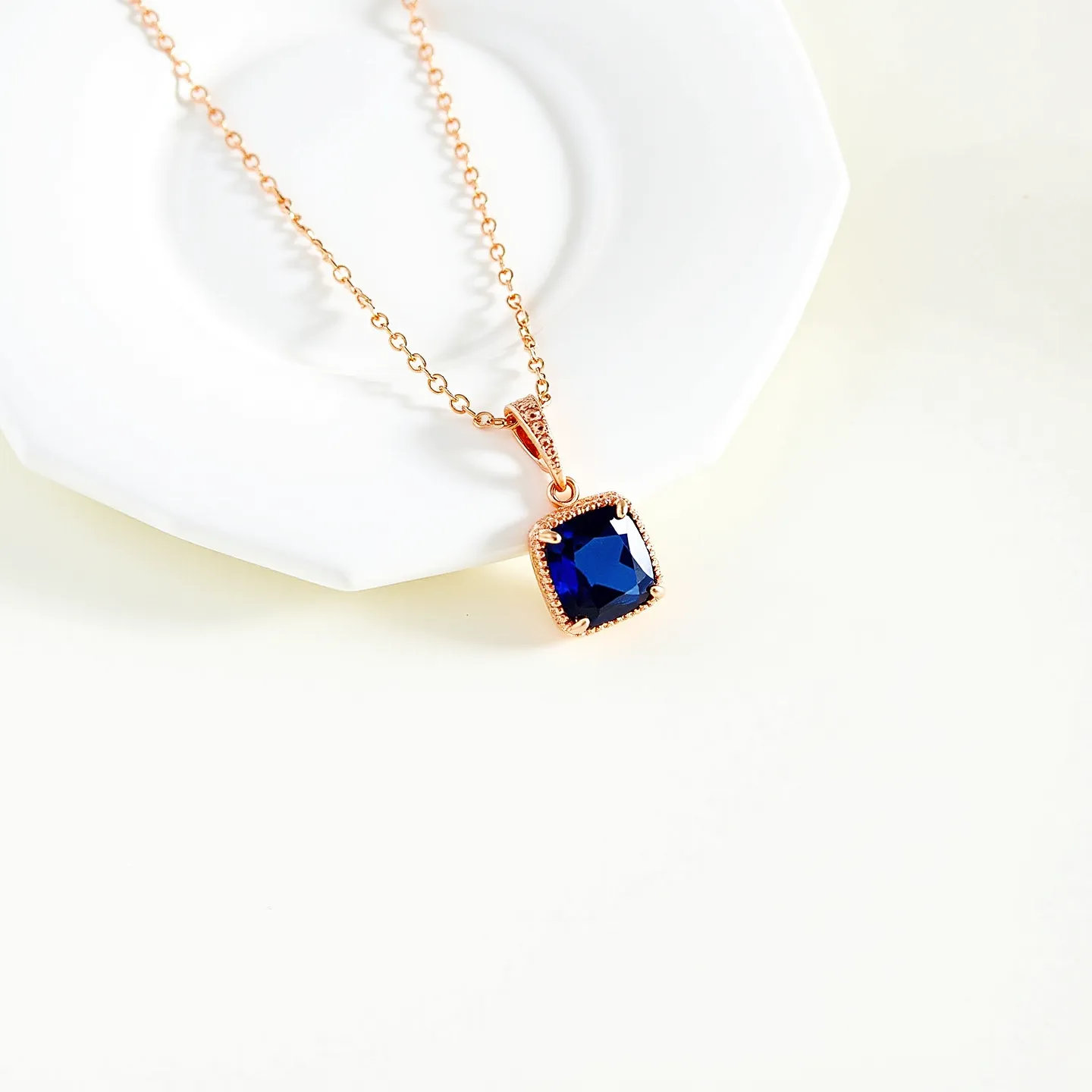 This sapphire necklace features a striking cushion-cut blue sapphire as its centerpiece, elegantly set in a rose gold bezel with a delicate milgrain border. The sapphire is securely mounted in the setting, which is further enhanced by a detailed bail connected to a fine chain, also made of rose gold. The chain consists of small, circular links, designed to complement the pendant while adding a seamless connection. The closure mechanism is not visible, but it is likely equipped with a discreet clasp that maintains the necklace's streamlined aesthetic.