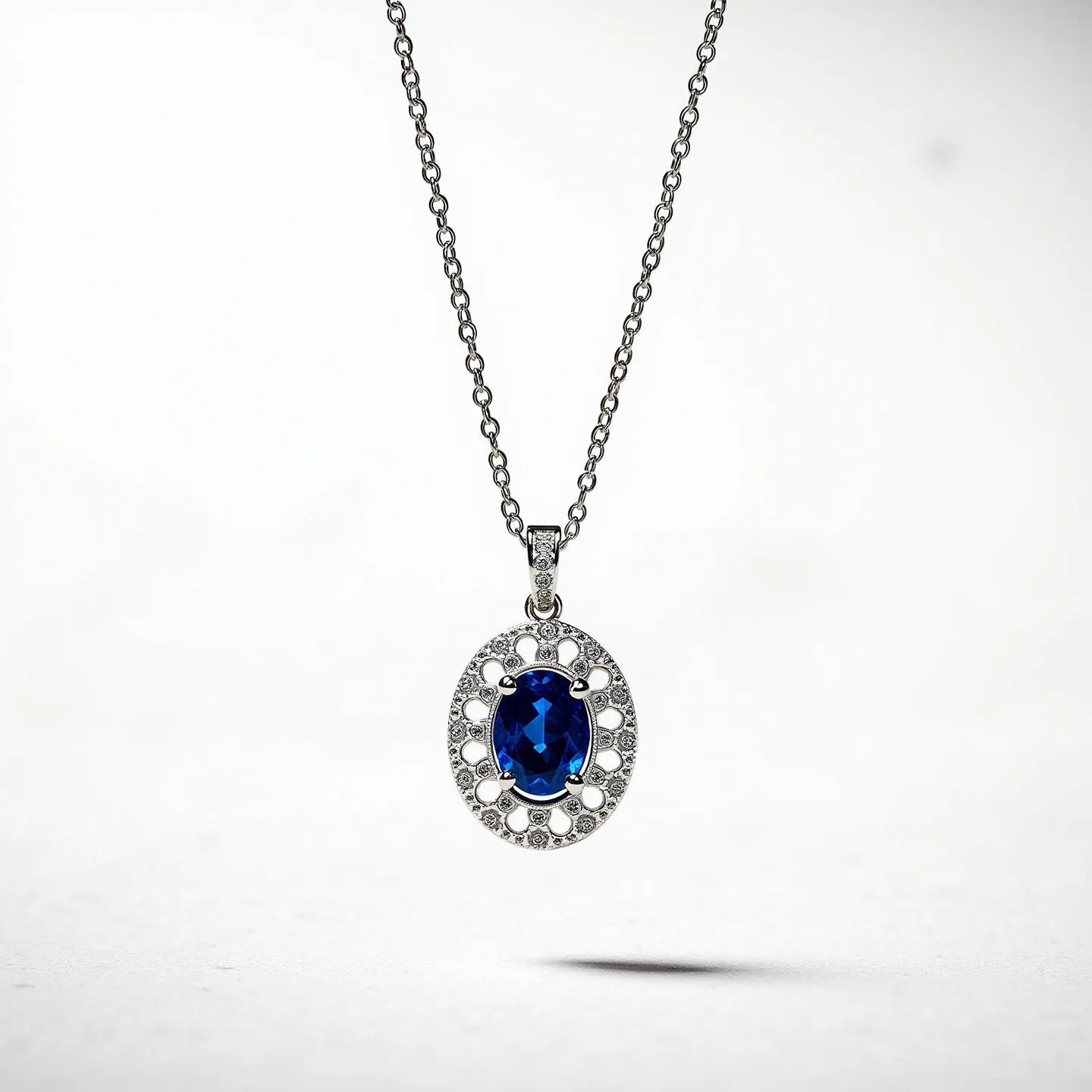 This sapphire necklace features an elegant design centered around a striking oval-cut sapphire gemstone, set in what appears to be a silver or white gold setting. The sapphire is surrounded by a halo of smaller round-cut diamonds or diamond-like stones, enhancing its brilliance and providing a striking contrast to the deep blue of the sapphire. The pendant is attached to a delicate chain, likely made of the same metal as the setting, with a classic linked design. The clasp is not visible, suggesting a continuous, seamless style that complements the overall refined aesthetic of the necklace.