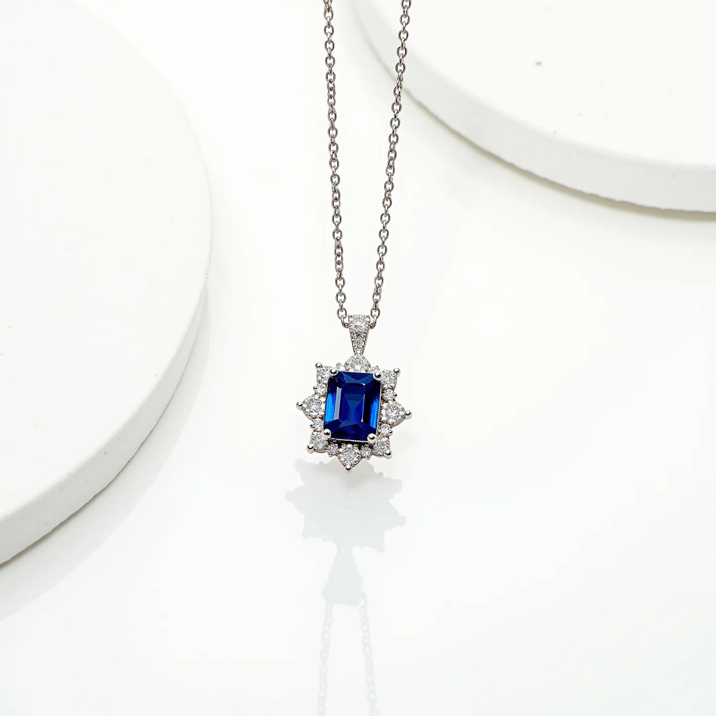 This sapphire necklace features a stunning rectangular-cut sapphire as its centerpiece. The deep blue stone is encircled by a halo of smaller round clear gems, which are likely diamonds, set in a symmetrical pattern that adds a touch of elegance and brilliance. The pendant is crafted in a bright metallic setting, possibly platinum or white gold, which complements the cool tones of the sapphire and accent stones. It hangs from a delicate chain that suggests sophistication and is secured with a standard clasp, ensuring both style and functionality.