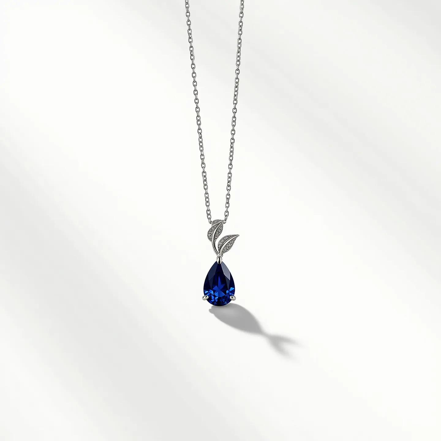 This sapphire necklace features a striking teardrop-shaped sapphire set in an elegant pendant design. The sapphire, with its deep blue hue, appears to be faceted, enhancing its brilliance and sparkle. It's beautifully set within a prong setting, which securely holds the gemstone while allowing maximum light to pass through. Above the sapphire are two leaf-like accents, adorned with smaller stones or intricate metalwork that add a delicate touch to the overall design. The necklace is completed with a fine metal chain, likely crafted from a precious metal such as white gold or platinum, known for its durable and lustrous qualities. The necklace is designed for secure wear, featuring a typical clasp mechanism for easy attachment and removal.