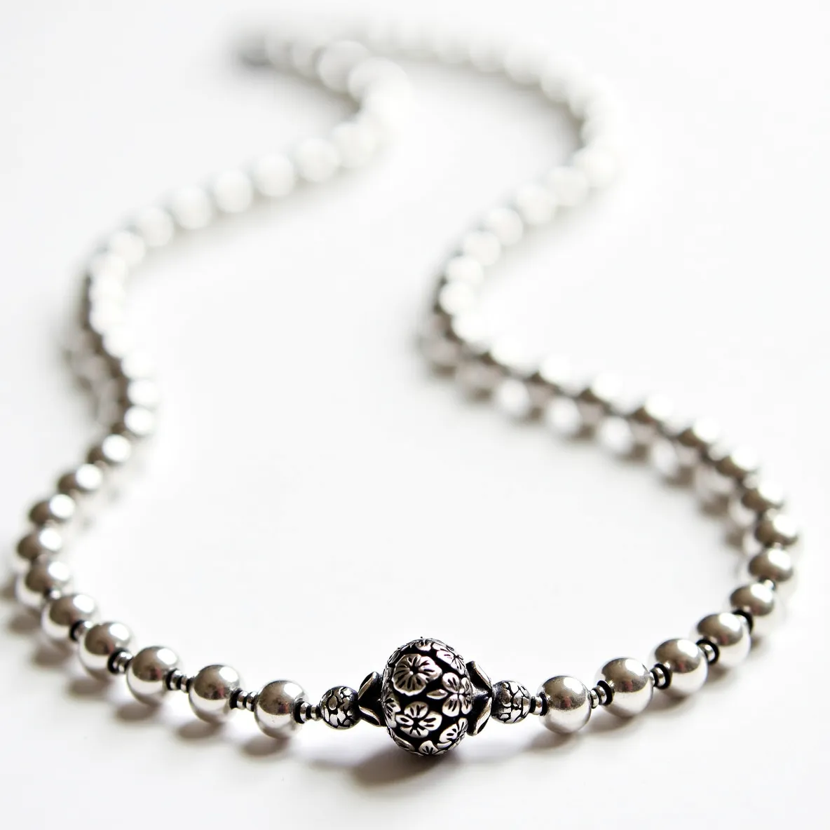 This silver bead necklace features a sequence of polished silver beads, each meticulously crafted to create a harmonious and elegant design. At the center, a larger focal bead with intricate floral engravings adds a touch of sophistication and artistry. The beads are uniformly spherical, enhancing the classic elegance of the piece. The necklace includes a discreet clasp that seamlessly integrates with the beads, ensuring both security and aesthetic continuity. The craftsmanship evident in the engraved center bead and overall uniformity highlights a blend of traditional and contemporary jewelry design.