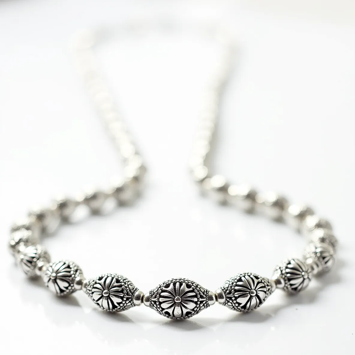 This silver bead necklace features intricately designed beads with floral motifs, showcasing a detailed and ornate pattern that suggests craftsmanship. The beads are likely crafted from sterling silver, given their reflective surface and intricate detailing. The necklace does not appear to include any additional gemstones or settings, focusing instead on the beauty of the silverwork. Each bead is spaced evenly along the strand, potentially strung on a durable metal or synthetic cord. The attachment or clasp is not visible in this particular view, suggesting a seamless or hidden clasp style that complements the overall aesthetic of the piece.