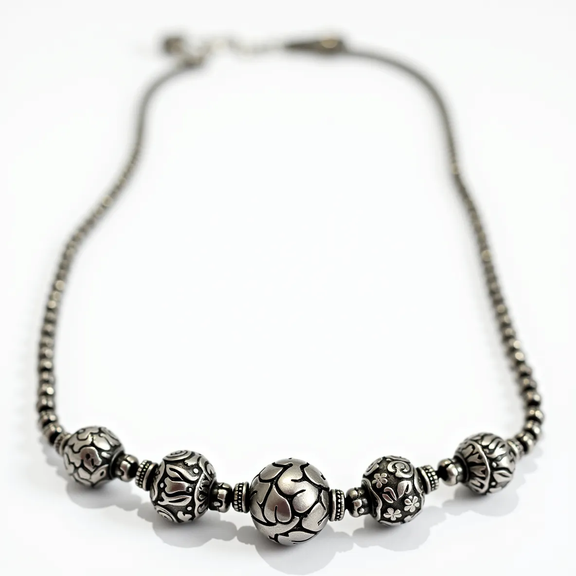 This silver bead necklace features intricately detailed beads, each showcasing ornate designs with a slightly oxidized finish that highlights the patterns. The beads are spherical, crafted from sterling silver, and vary in size, creating a visually appealing graduated effect along the necklace. The central bead is the largest, surrounded by smaller, equally detailed beads, adding depth and texture to the piece. The necklace is equipped with a lobster clasp attachment, ensuring secure wearability. This design blends traditional craftsmanship with contemporary styling, making it a versatile accessory for various occasions.