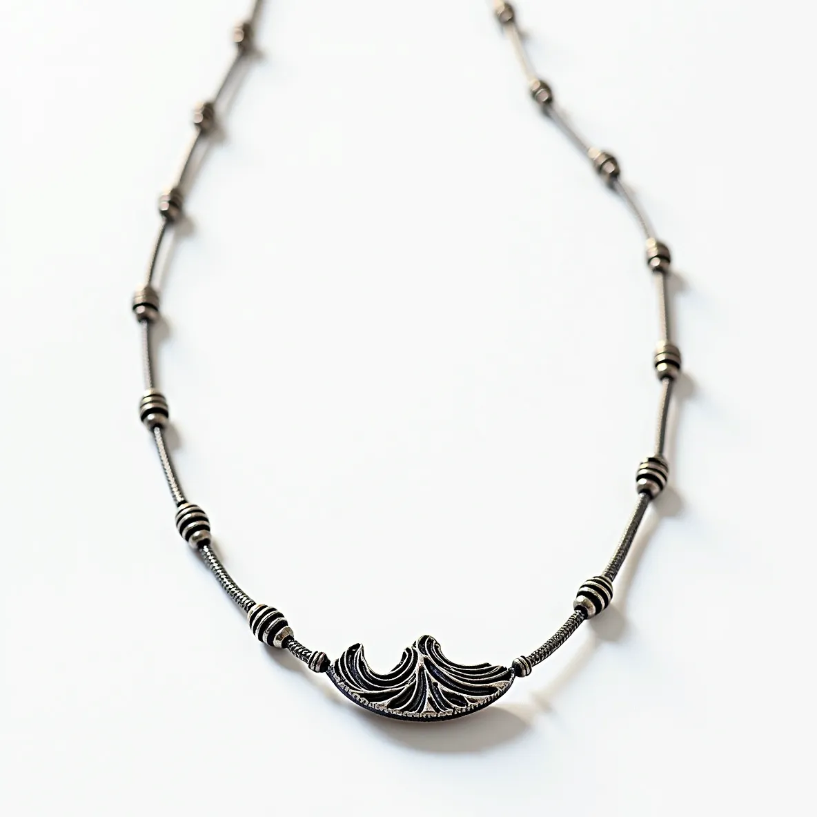 This silver bead necklace features a series of intricately detailed silver beads, each separated by sections of smooth, flexible silver chain. The central focal point is a uniquely designed, ornate silver element with engraved patterns, adding a touch of elegance to the piece. The beads are uniform in shape and size, providing a balanced and rhythmic aesthetic. The necklace is constructed entirely of silver, offering a timeless, metallic sheen. It is fastened with a simple yet secure clasp, ensuring both functionality and style. The overall design exudes a sophisticated and classic appeal, perfect for various occasions.