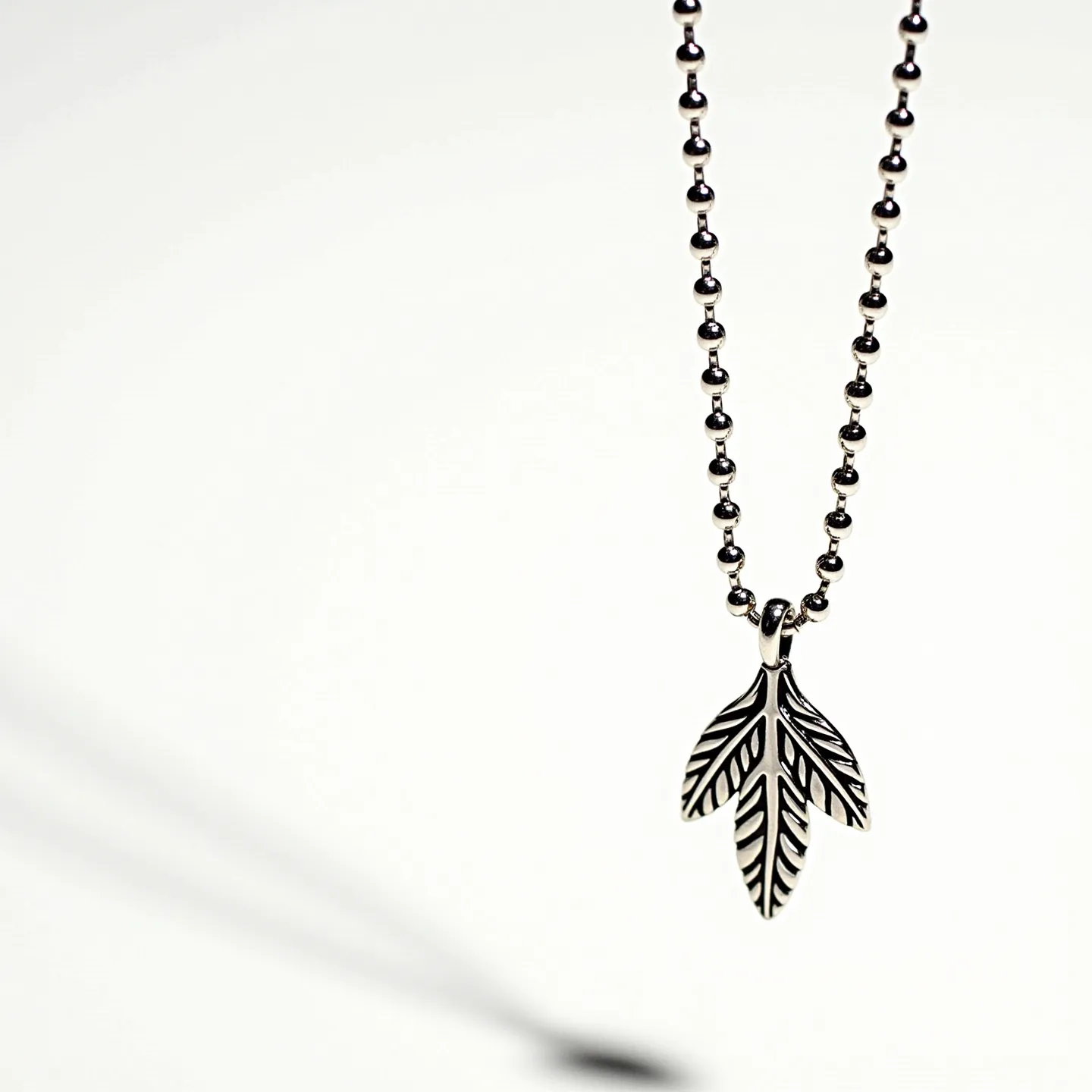 This silver bead necklace features a series of small, polished beads strung together to create a continuous chain. The necklace is adorned with a pendant in the shape of a stylized leaf, adding an artistic touch to its design. The pendant is intricately detailed with engraved patterns that mimic the veins found on a real leaf, enhancing its visual appeal. The necklace most likely includes a simple ball chain clasp, allowing for easy attachment and removal. The absence of any gemstones focuses attention on the elegant simplicity and craftsmanship of the silver design elements.