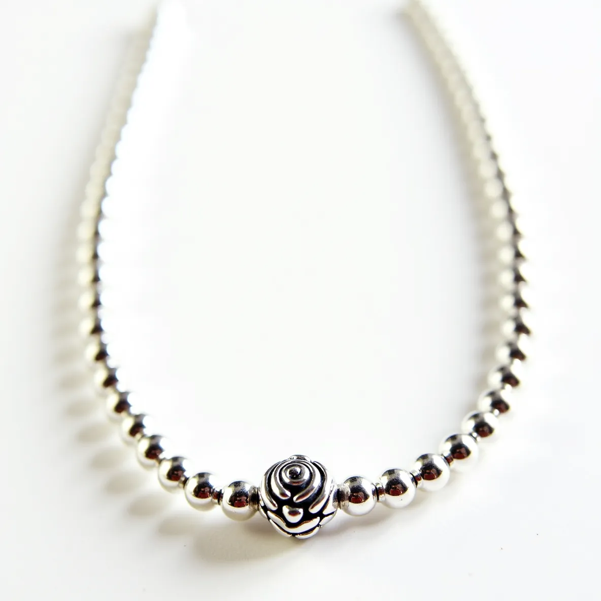 This silver bead necklace features smooth, polished beads that are uniform in size and strung together in an elegant sequence. At the center, there is a distinctive bead crafted in the shape of a rose, with intricate detailing that adds a focal point to the piece. The necklace is likely constructed from sterling silver, providing a gleaming finish that highlights the craftsmanship. A secure clasp is attached, ensuring easy wearability and removal, though the specific type of clasp is not detailed in this view. This design combines simplicity with a touch of floral elegance, making it a versatile accessory for various occasions.