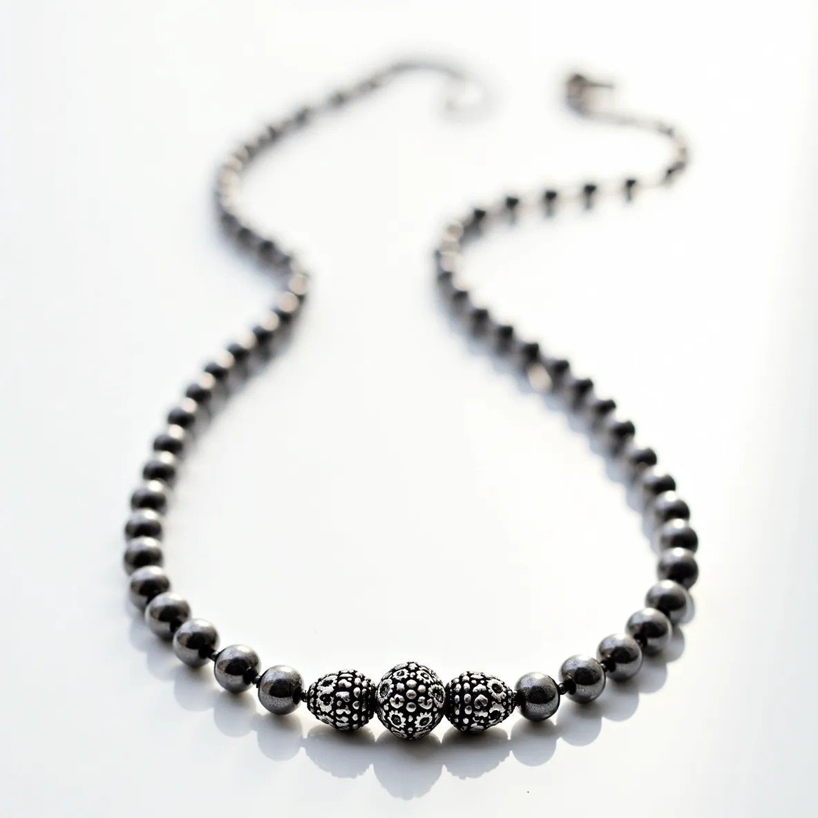 This silver bead necklace features a series of smooth, polished silver beads strung together, creating a sleek and continuous line. At the center, there are three distinctively larger beads with ornate, decorative patterns, adding a touch of intricacy and elegance. These central beads appear to be crafted from the same silver material, designed with embossed or engraved floral motifs. The necklace is finished with a simple hook-and-eye clasp, providing a secure and seamless closure. The overall design is balanced and graceful, combining both simplicity and detailed craftsmanship.