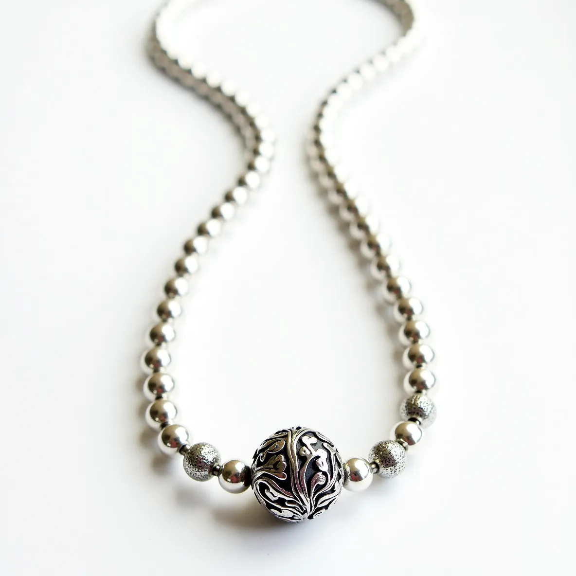 This silver bead necklace features a series of polished, spherical silver beads, creating a uniform and elegant appearance. At the center, a larger bead stands out with intricate engravings, adding a touch of intricacy and sophistication. The addition of several textured silver beads near the central feature enhances the contrast and visual interest. The necklace is completed with a silver clasp, ensuring secure fastening while maintaining a cohesive metallic aesthetic. The use of high-quality silver in both the beads and the clasp highlights the craftsmanship and attention to detail in this elegant piece.