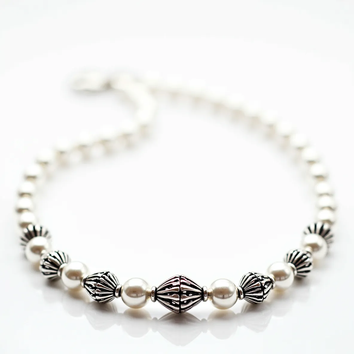 This silver bead necklace features a combination of smooth, spherical white beads, which may be faux pearls, interspersed with ornate silver beads. The silver beads display intricate designs, likely crafted from sterling silver, providing a vintage aesthetic. The central silver bead is larger and uniquely shaped, emphasizing the necklace's focal point. A standard, secure clasp is present, ensuring the necklace stays in place when worn. The symmetry and alternating pattern of silver and white beads create a balanced and elegant design.