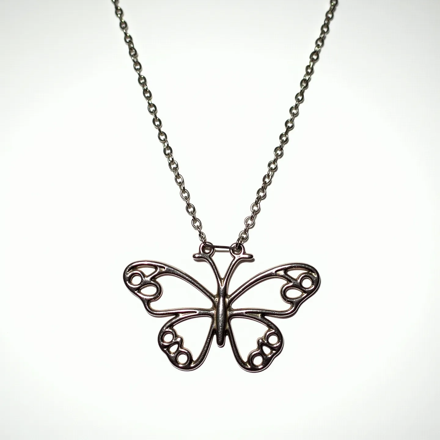 This silver butterfly necklace features an intricately designed pendant with delicate openwork detailing, creating an elegant representation of a butterfly. The pendant is crafted from silver, showcasing a polished finish that highlights its graceful curves and symmetrical design. The necklace is suspended from a fine silver chain, which complements the pendant's refined aesthetic. The attachment appears to be seamless, providing an uninterrupted flow between the pendant and the chain. A simple clasp, likely a spring ring or lobster claw, secures the necklace, ensuring ease of wear and durability.