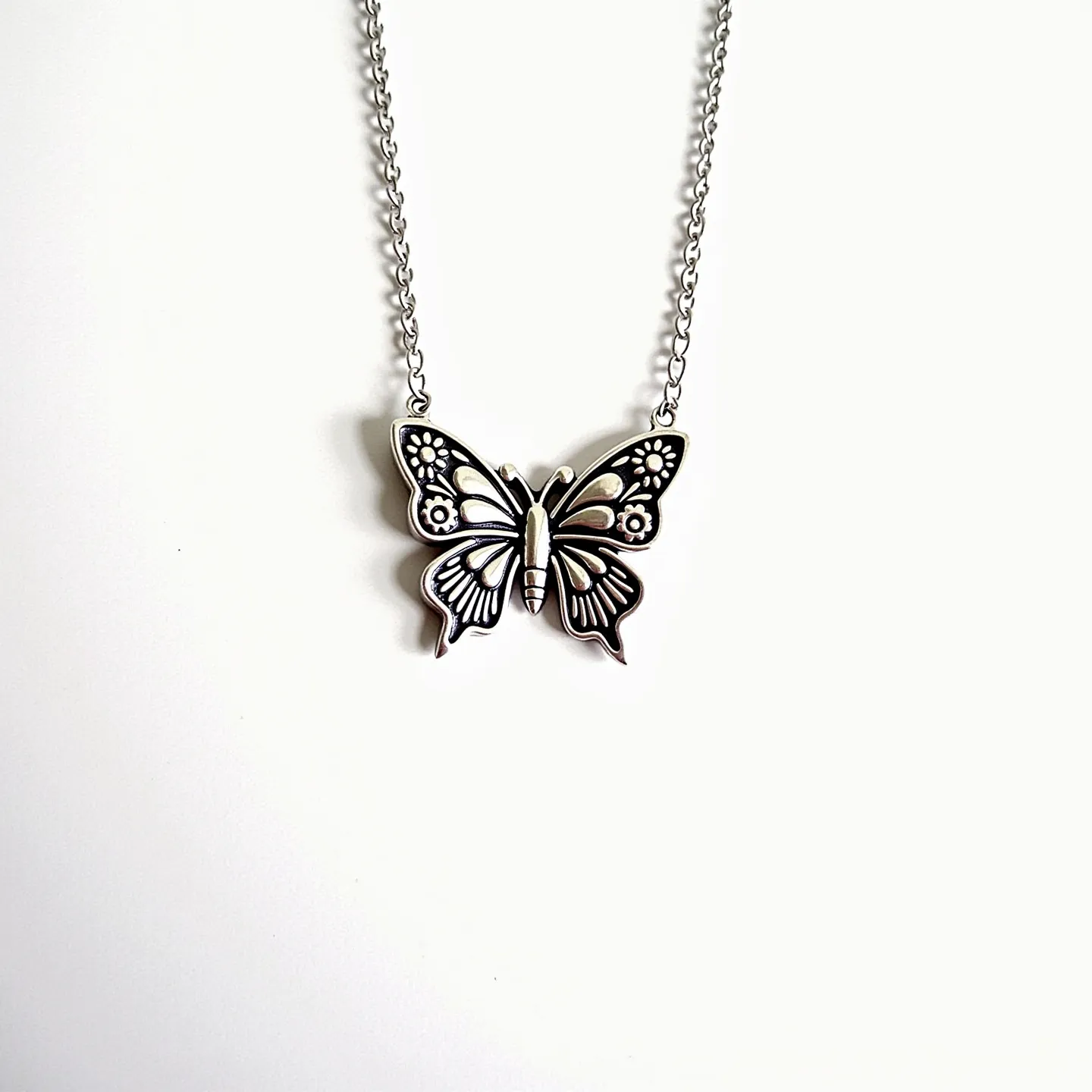 This silver butterfly necklace features a beautifully crafted butterfly pendant made of silver, showcasing intricate detailing on the wings, which includes floral and linear patterns for an added decorative touch. The chain is composed of small, evenly linked silver loops, providing a delicate and elegant look that complements the pendant. The necklace has a simple clasp designed for ease of use while ensuring secure attachment, seamlessly blending functionality with its aesthetic design.