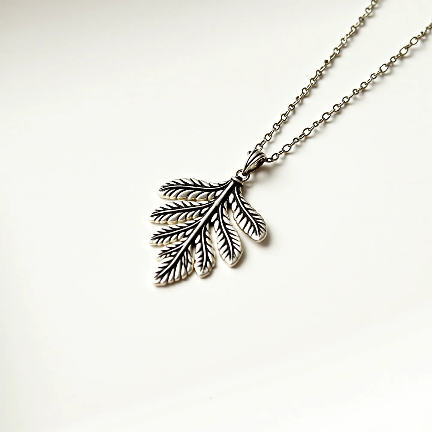 This silver chain features an elegant, intricate pendant shaped like a cluster of leaves. The pendant is expertly crafted with detailed vein patterns that enhance its organic aesthetic. The chain itself consists of small, round links that give it a delicate yet sturdy appearance. There are no gemstones or additional embellishments on the pendant or chain, allowing the detailed leaf design to be the focal point. The pendant is attached seamlessly to the chain with a simple loop, ensuring it hangs securely and with fluidity. The overall design is both sophisticated and nature-inspired.