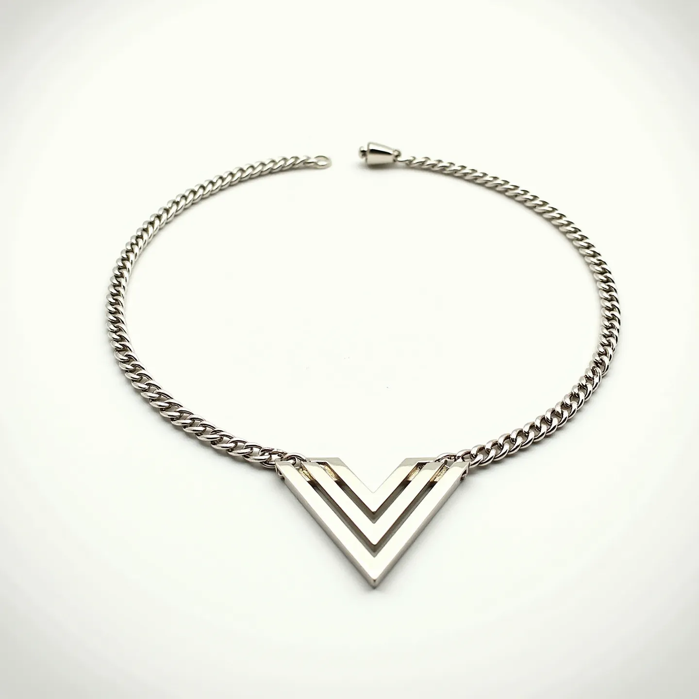 This silver chain features a polished, interlocking curb link design, exuding a sleek and modern aesthetic. At its center, a geometric V-shaped pendant crafted from the same silver material adds a striking focal point. The pendant showcases a layered design, providing depth and visual interest without any gemstones or additional embellishments. The entire piece is secured with a lobster clasp, ensuring a secure and adjustable fit for the wearer. The simplicity and elegance of the chain and pendant make it a versatile accessory suitable for various styles and occasions.