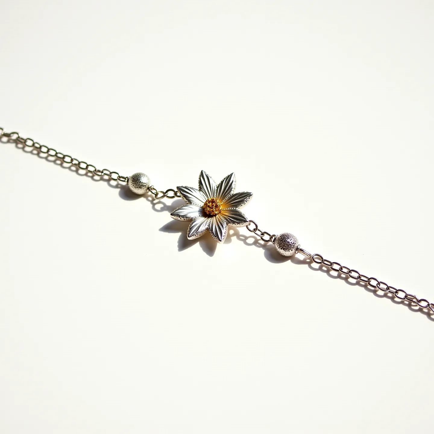 This silver chain features a delicate metal construction, accented by decorative silver spheres. At its center is a floral motif with intricately crafted petals, and at the heart of this flower lies a vibrant orange gem, cut in a round shape. The gem is securely set, adding a splash of color to the overall design. The chain includes standard links, facilitating flexibility and complementing the ornamental centerpiece. The attachment style is not visible in this view, ensuring a seamless look that highlights the artistic elements of the piece.