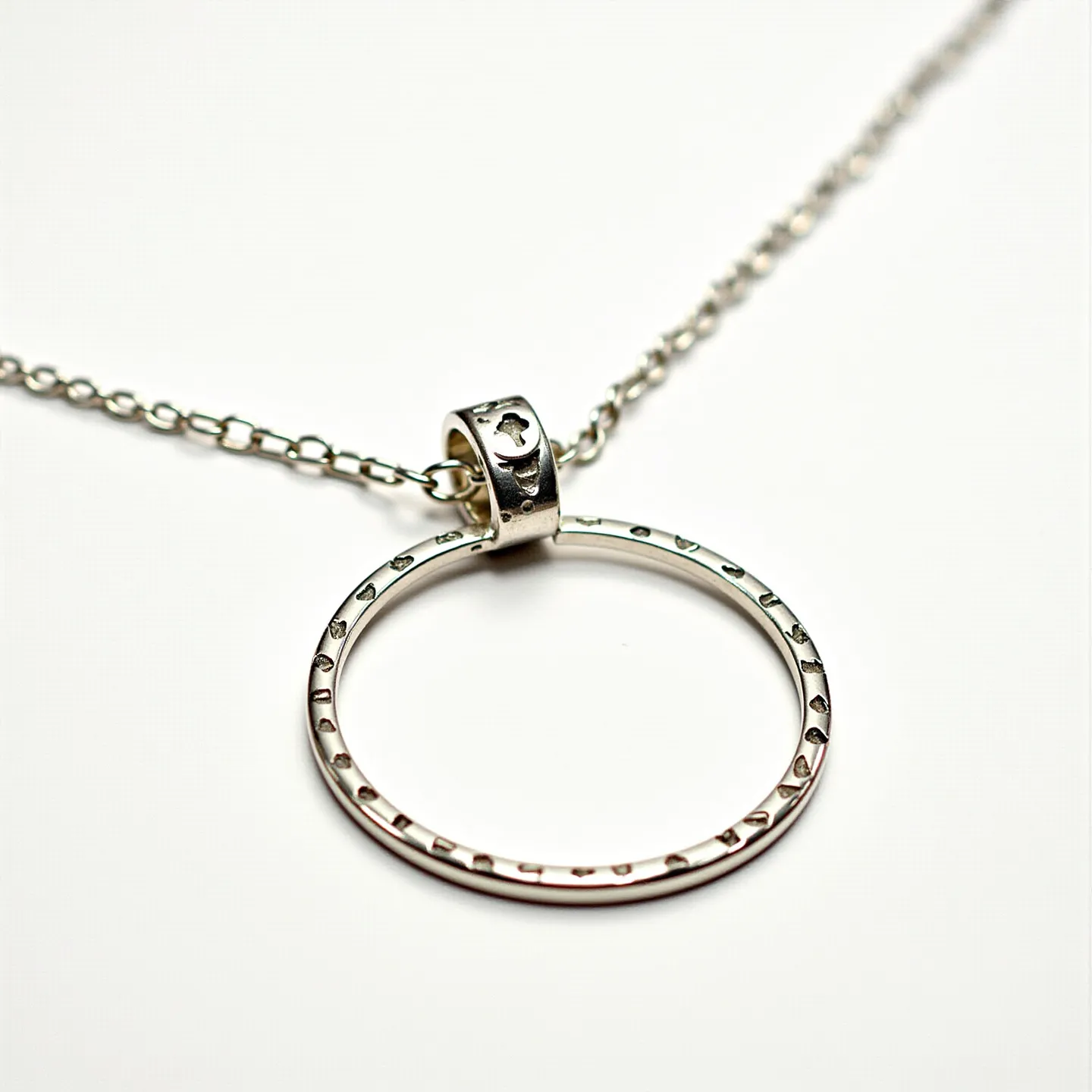 This silver chain features a delicate, polished ring pendant as its central element. The pendant is embellished with small engraved patterns along its surface, adding subtle detail and texture. The chain itself is of a classic linked design, providing both elegance and durability. The pendant is attached to the chain via a small loop, which is also adorned with engraved details that complement the design of the pendant. This craftsmanship highlights the simple yet sophisticated aesthetic of the piece.