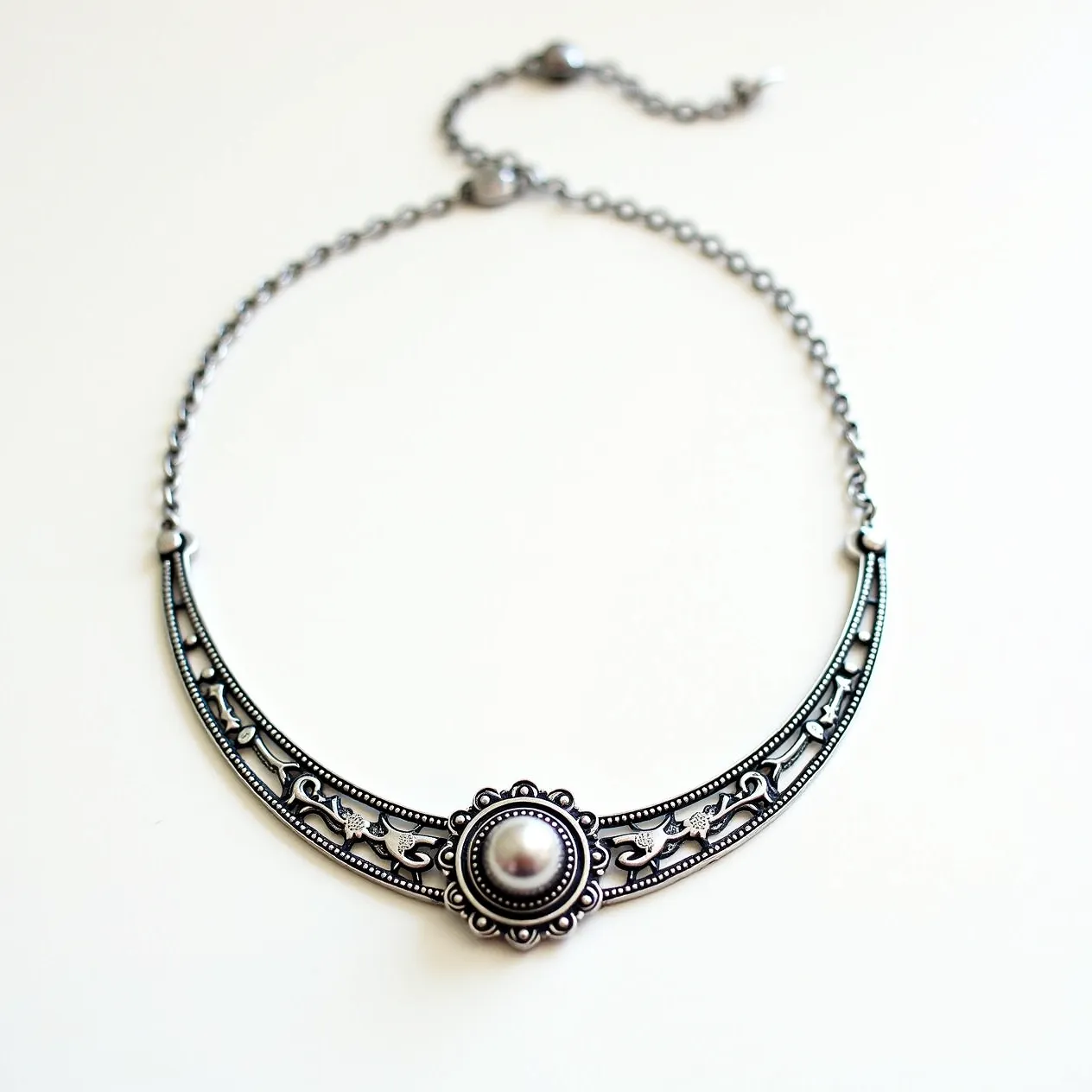 This silver choker necklace features an ornate design with an intricate filigree pattern embellished with floral and vine motifs. The centerpiece is a round, pearl-like gem set in a bezel setting, adorned with a decorative border of small beaded details. The necklace includes a delicate chain that connects to the main decorative piece and is closed with a simple lobster clasp for secure attachment. The combination of silver metalwork and the central gem creates an elegant and timeless accessory.