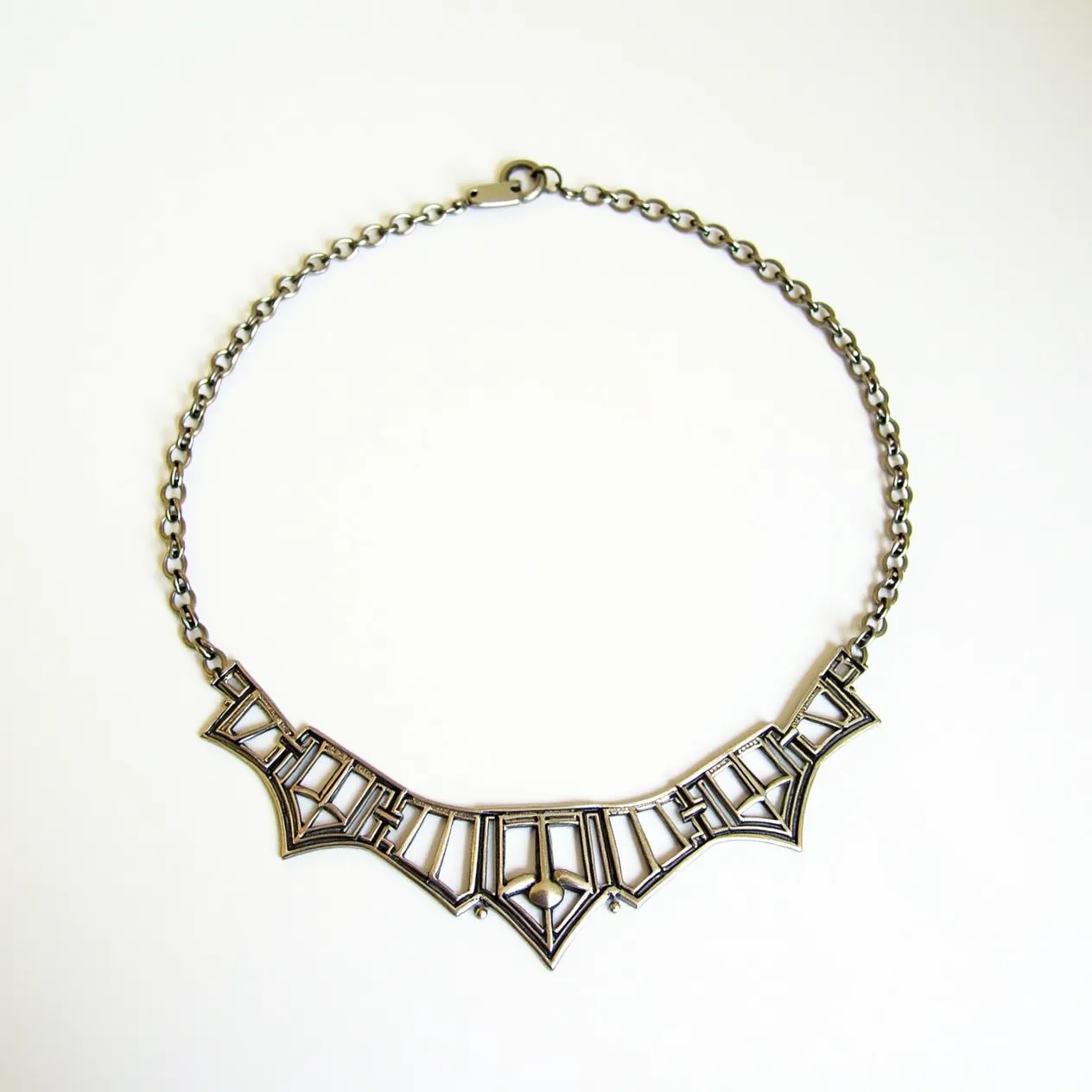 This silver choker necklace features an intricate geometric design that stands out with bold, angular shapes. The necklace is crafted from silver, giving it a sleek, polished appearance. The front of the choker showcases a series of interconnected, angular patterns that resemble a stylized interpretation of art deco influences. The choker is linked to a chain made of matching silver loops, providing a cohesive look throughout. For closure, the necklace has a simple hook clasp, ensuring a secure fit when worn. The minimalist yet striking design makes it a versatile accessory suitable for a variety of occasions.