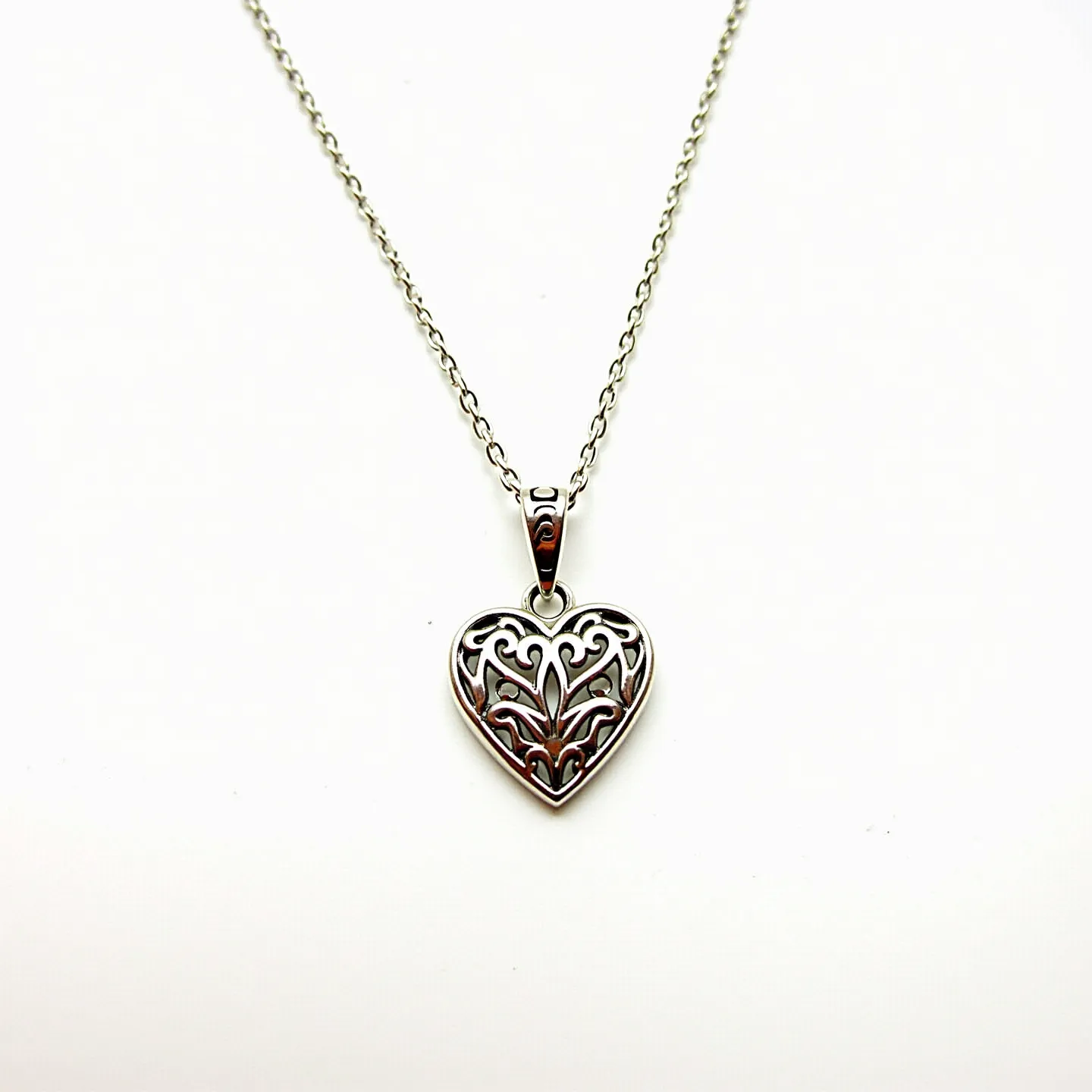 This silver heart necklace features an intricately designed heart-shaped pendant, crafted from a polished silver material. The pendant displays an elegant filigree pattern, adding a touch of artistry and sophistication to its overall design. The chain, characterized by small, interlinked loops, enhances the delicate appearance of the piece. A simple yet secure bail connects the heart pendant to the chain, ensuring that it hangs gracefully. The necklace is likely fastened with a standard clasp, providing both security and ease of wear.