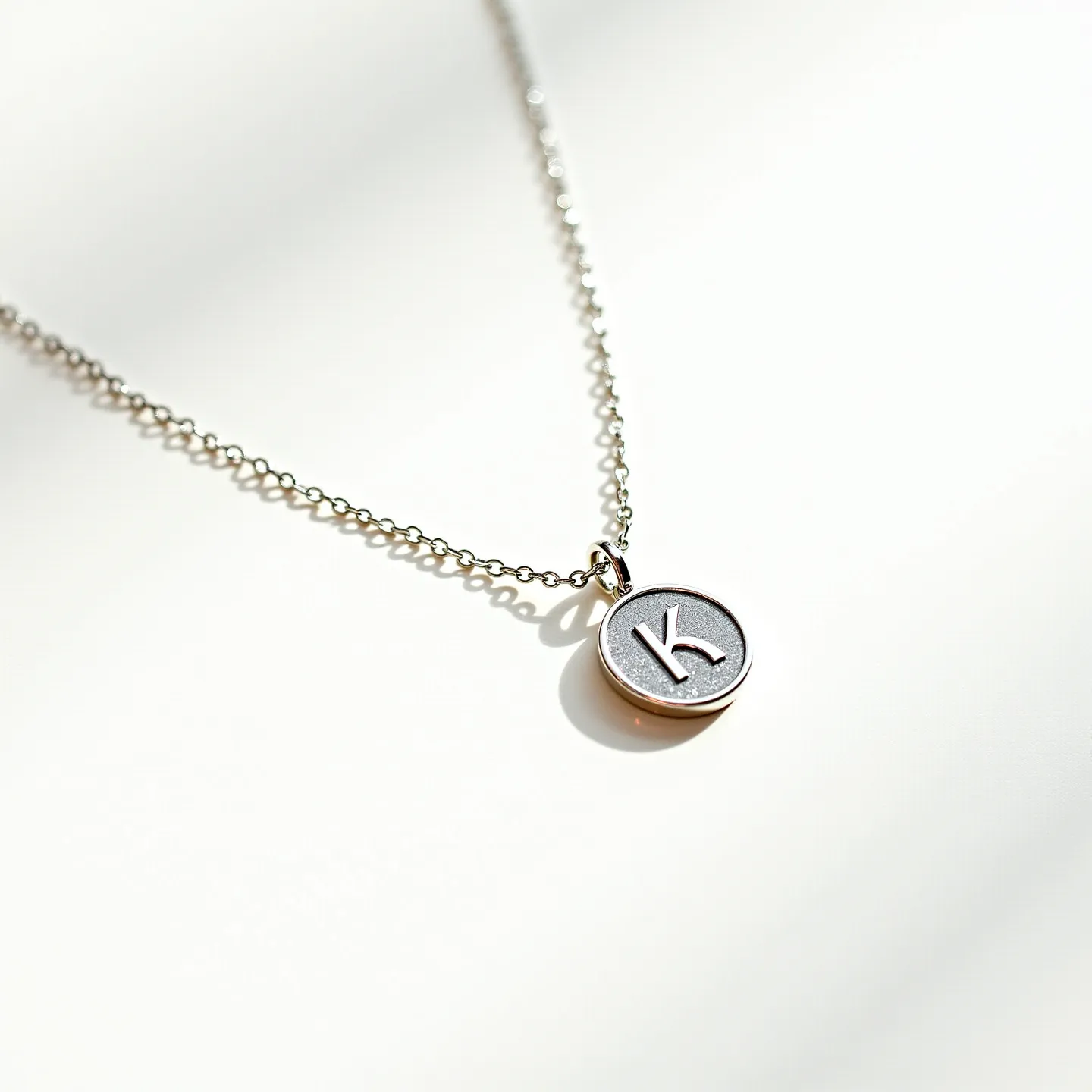 This silver initial necklace features a delicate chain crafted from sterling silver, which supports a pendant with a raised letter "K" inscribed on a circular disc. The pendant has a polished finish with a textured background, providing a subtle contrast to the shiny letter. The simple yet elegant design of the chain is characterized by small, connected links, offering a dainty appearance. The clasp appears to be a typical lobster claw design, ensuring secure attachment while allowing for easy opening and closing.