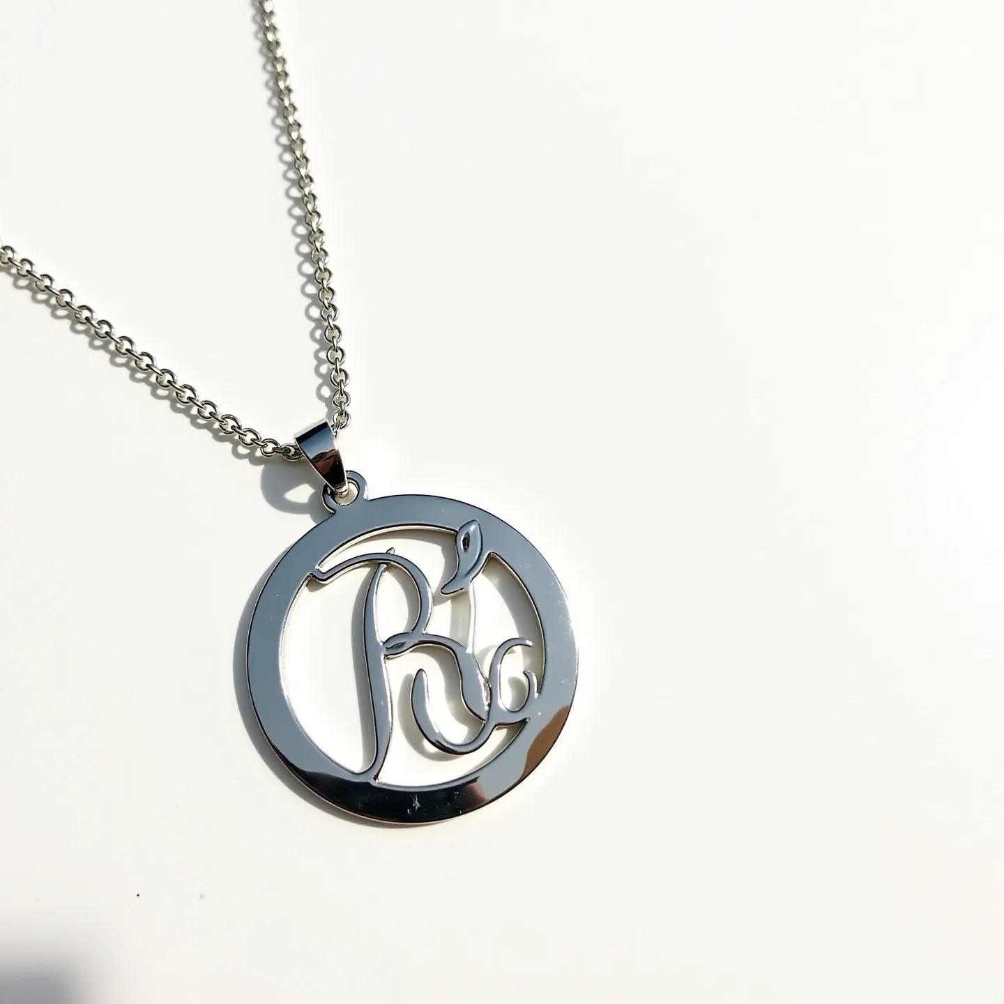 This silver initial necklace features a circular pendant with a polished, reflective surface, prominently displaying the intricate script of the initial "R" within it. The pendant is crafted from high-quality silver, exhibiting a smooth and lustrous finish, which adds a touch of elegance to the design. It is suspended from a delicate yet sturdy chain, also crafted from silver, which complements the pendant's shine. The connection between the pendant and chain is facilitated by a sleek bail that ensures seamless movement along the chain. The necklace is secured with a classic clasp, likely a lobster or spring ring, ensuring both reliability and ease of use for the wearer.