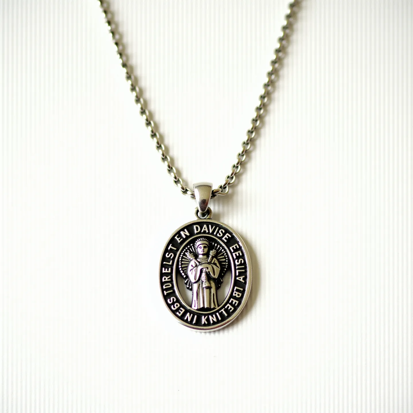 This St. Jude necklace features an oval pendant with a detailed depiction of St. Jude in the center. The pendant is crafted from a silver-toned metal, giving it a polished and durable appearance. Surrounding the figure is an engraved border with text, adding a structured and meaningful design element. The pendant hangs from a sturdy, matching chain that enhances the overall metallic sheen of the piece. The attachment is a sleek, elongated bail that securely connects the pendant to the chain, creating a seamless and cohesive look that is both functional and stylish. The clasp used is a straightforward lobster clasp, known for its reliability and ease of use.