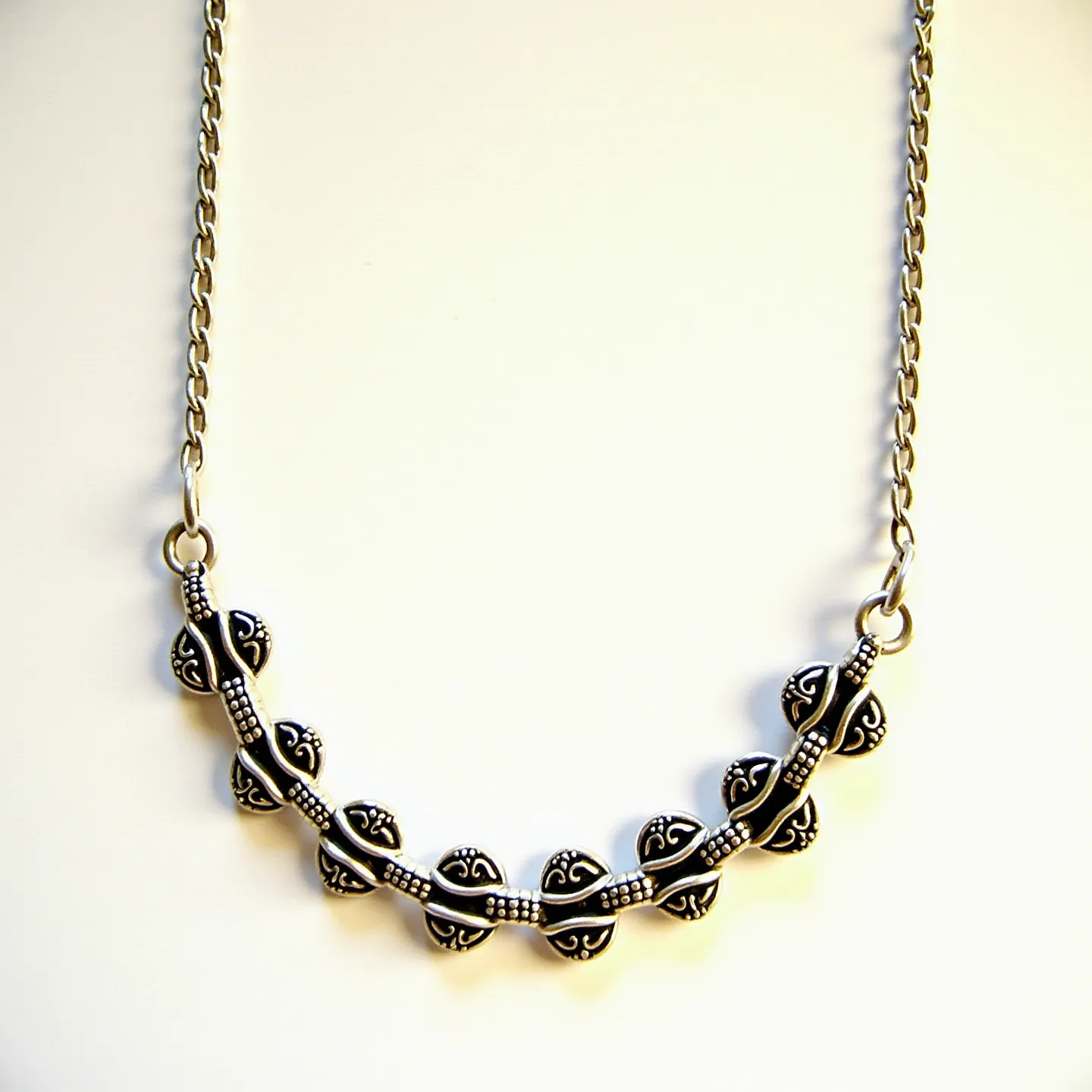 This sterling silver necklace features a chain with an intricate pendant design composed of decorative elements resembling small, rounded shapes with engraved patterns. The pendant elements are connected by a series of small, linked bars that create a harmonious and symmetrical appearance. The necklace has two circular links at each end that suggest a seamless integration with the chain, indicating a continuous flow of design. No gemstones or additional materials are present, highlighting the elegance of the silverwork. The necklace likely includes a conventional clasp mechanism for fastening.