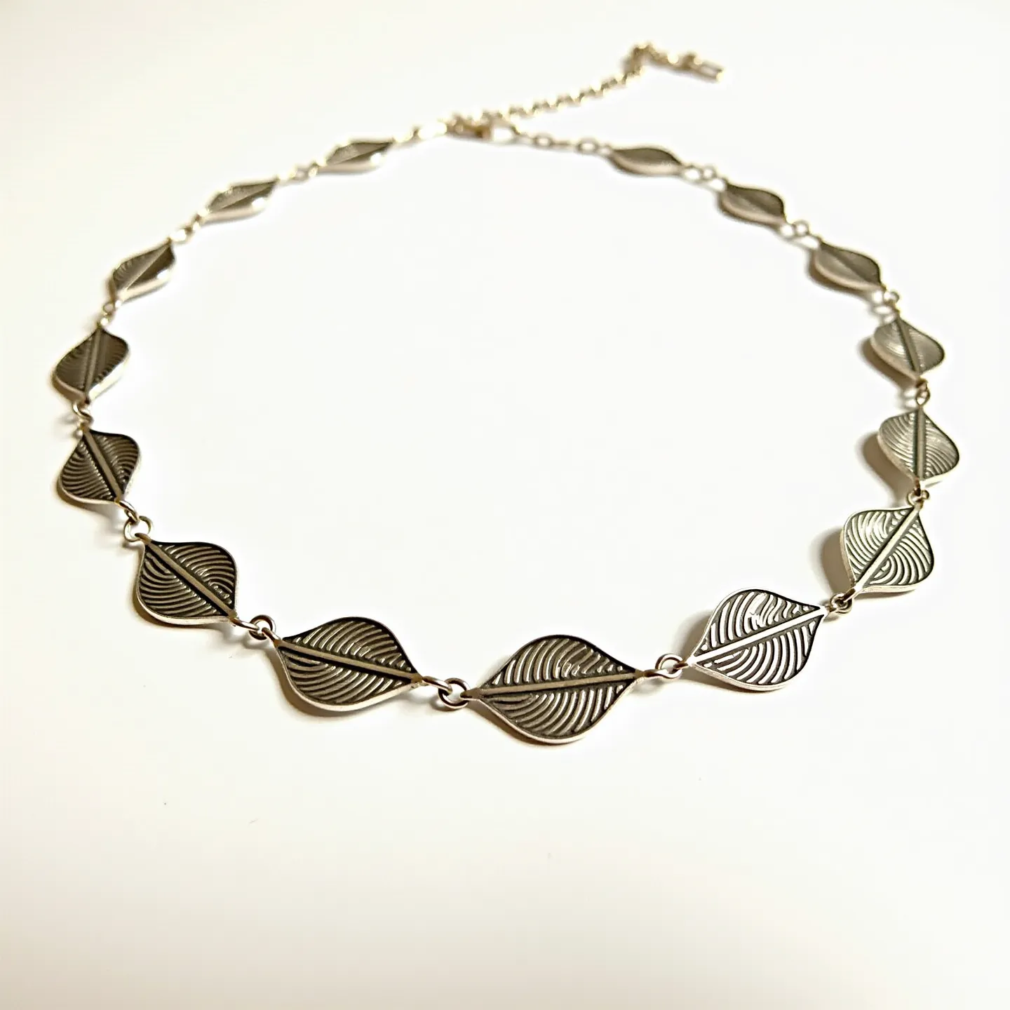 This sterling silver necklace features a series of intricately designed leaf-shaped links that are beautifully interconnected. The silver leaves are detailed with fine lines to mimic the natural veining found in real leaves, offering a textured and elegant appearance. The necklace is secured with a standard lobster clasp, accompanied by an adjustable chain extension to ensure a comfortable fit. The overall design showcases a delicate yet striking aesthetic, perfect for a nature-inspired look.