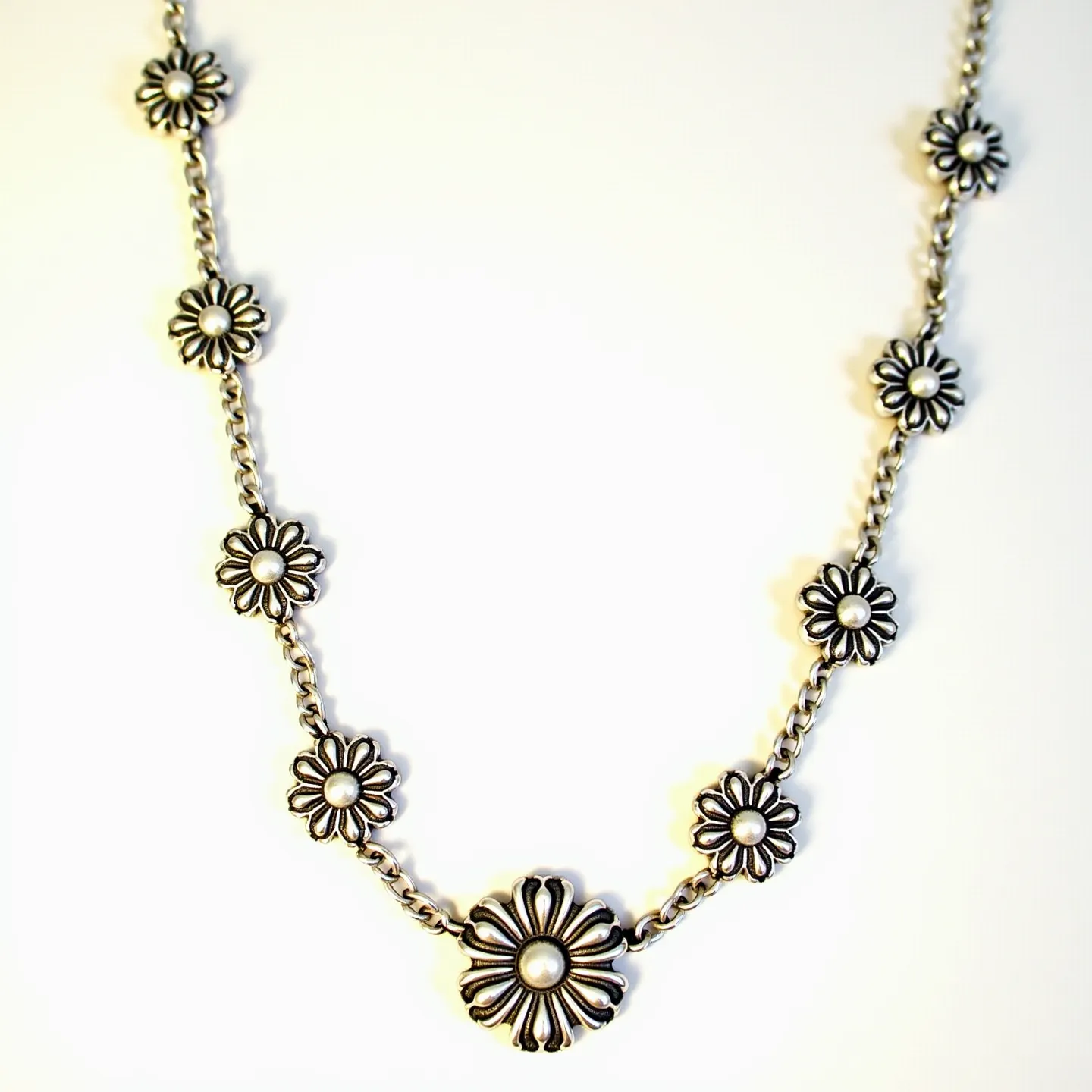 This sterling silver necklace features a series of intricate flower motifs, each with a raised, rounded center suggestive of a small, polished gem or bead, though its exact material is undetermined. The floral elements are evenly spaced along a chain link, enhancing the necklace's decorative appeal. The central flower is slightly larger, serving as a focal point in the design. This piece likely includes a standard clasp for securing the necklace, integrating seamlessly with the silver chain to ensure both functionality and aesthetic cohesion.